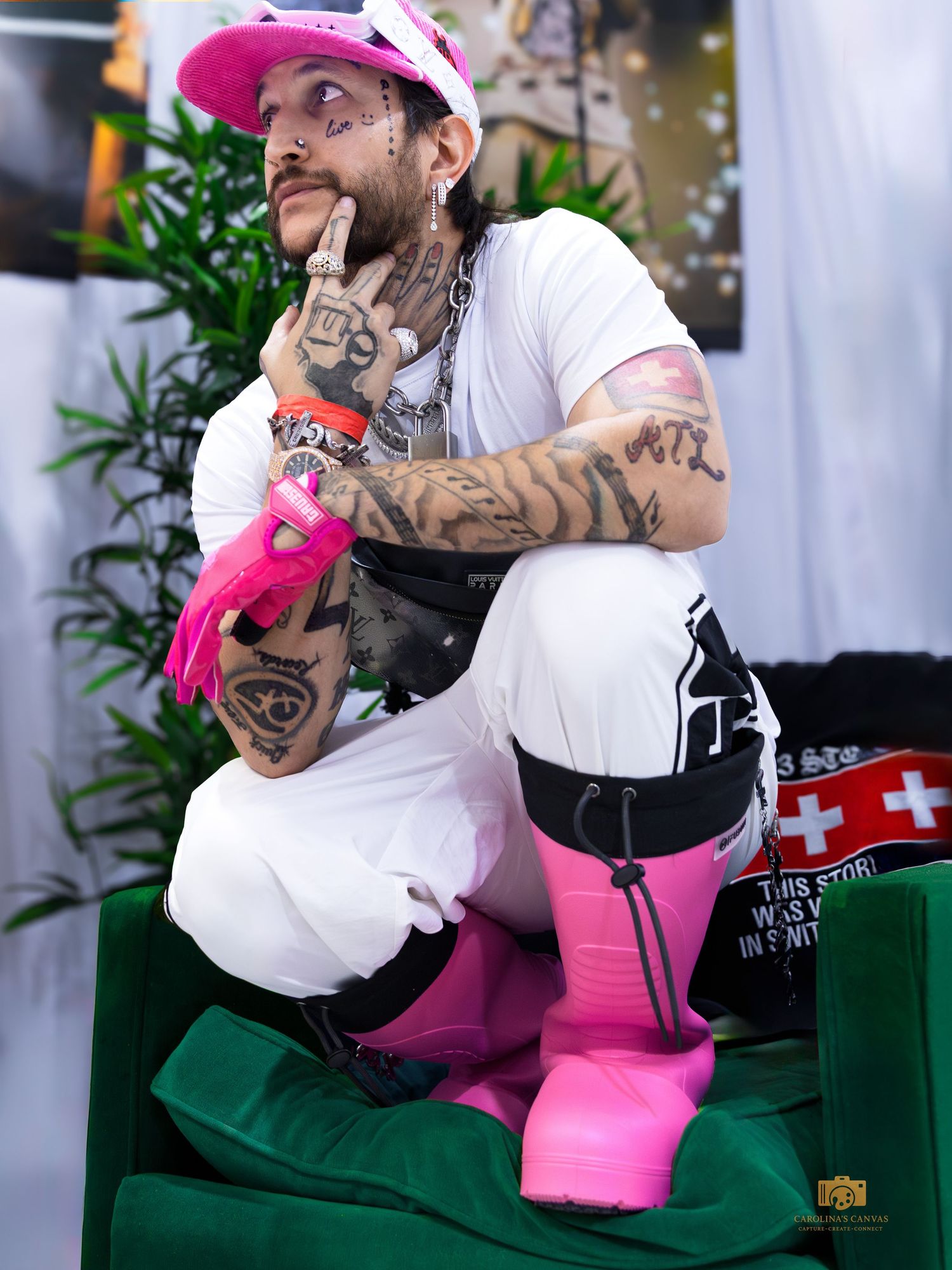Heavily tattooed man in white outfit with pink boots, gloves, and cap sitting pensively.