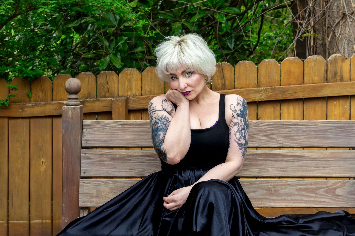 Woman with blonde hair and tattoos wearing a black dress seated on a wooden bench outdoors.