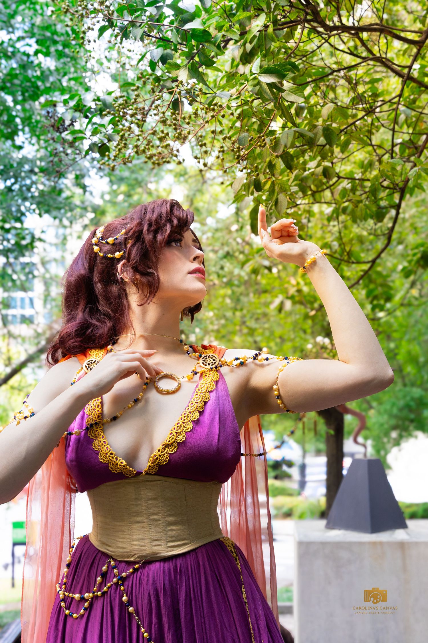 Woman in purple and gold fantasy costume poses gracefully under green tree branches outdoors.