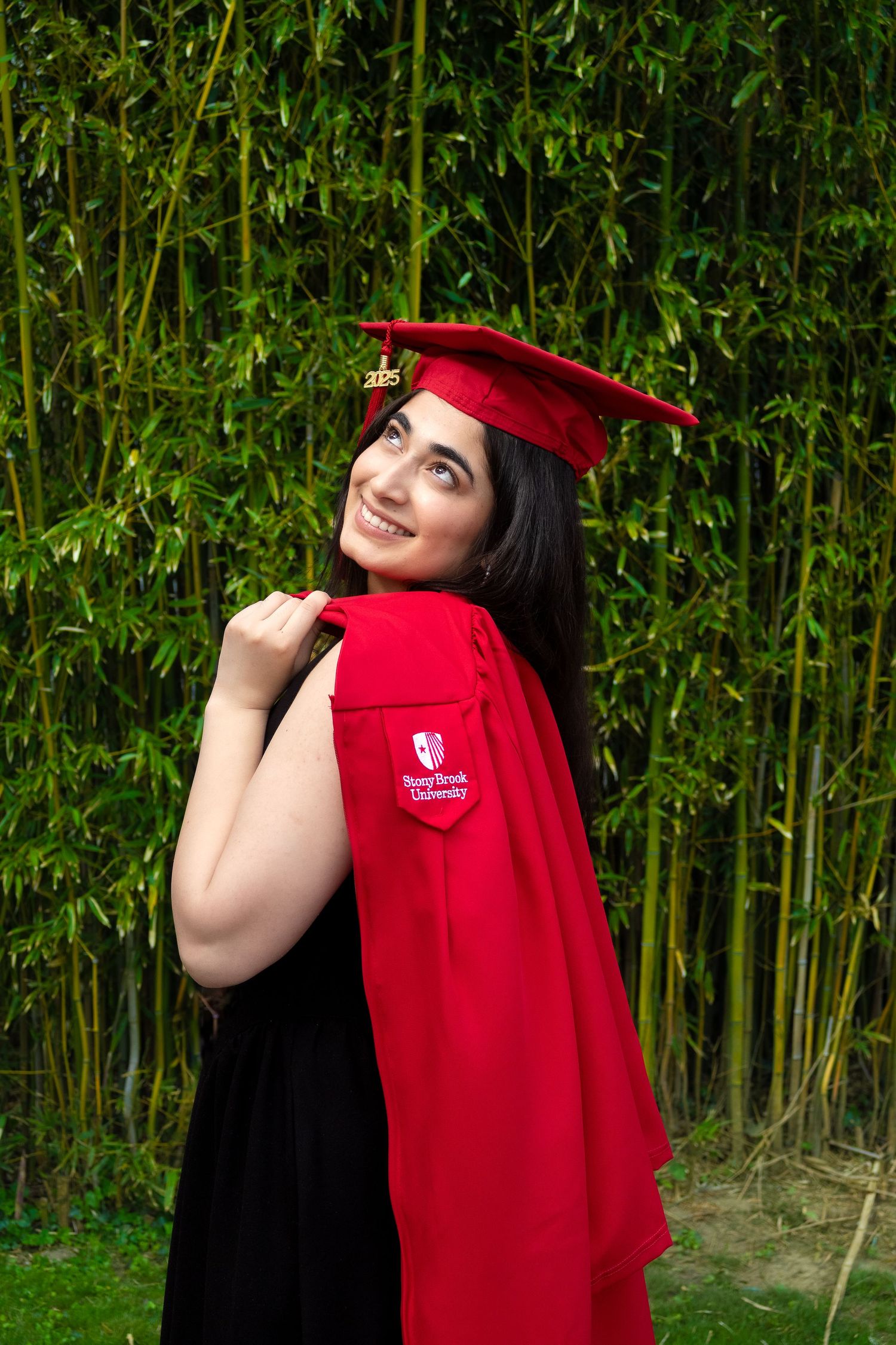 A graduate in red cap and gown poses cheerfully against a green bamboo wall backdrop.