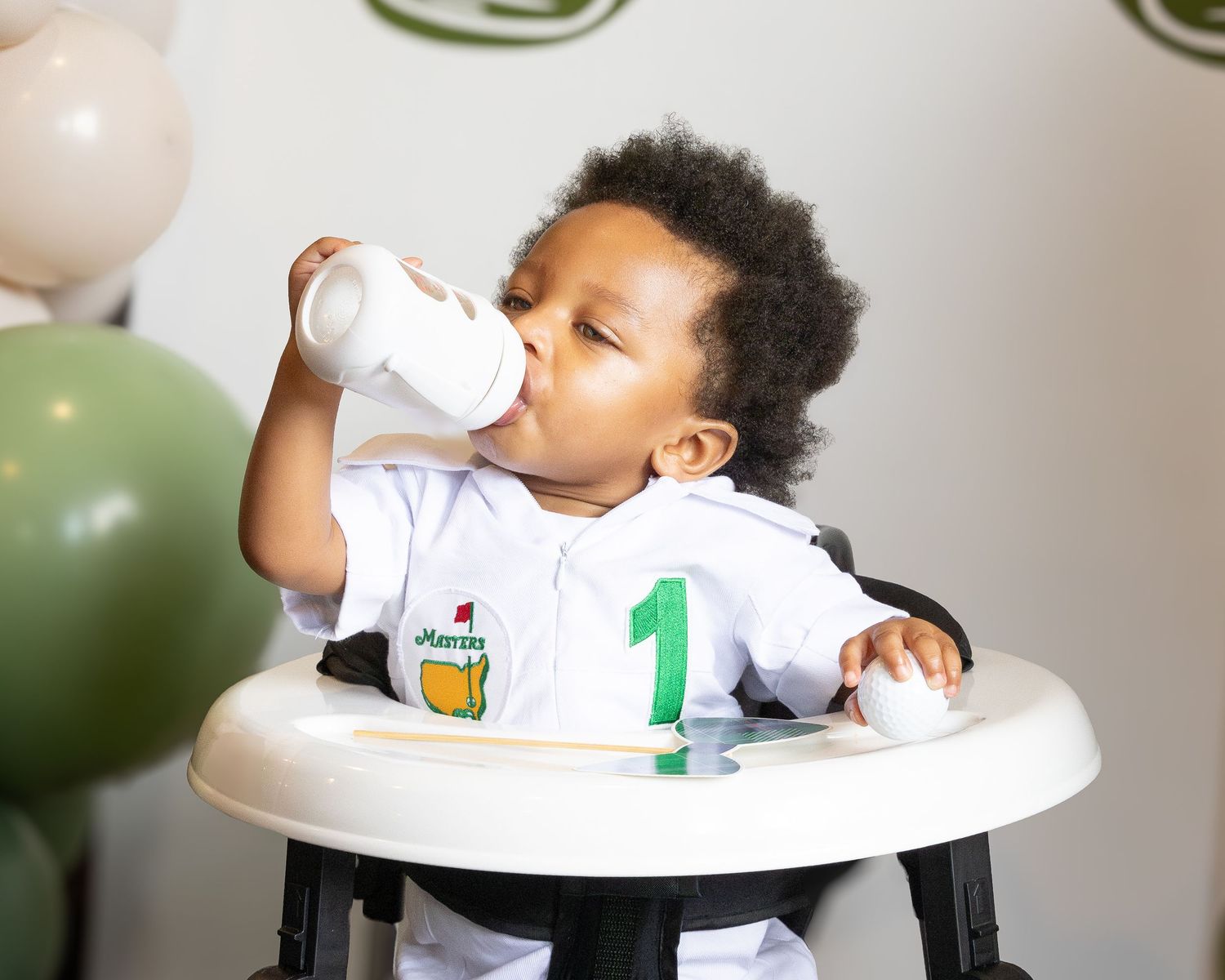 Baby in white shirt sits in high chair drinking from bottle during feeding time.