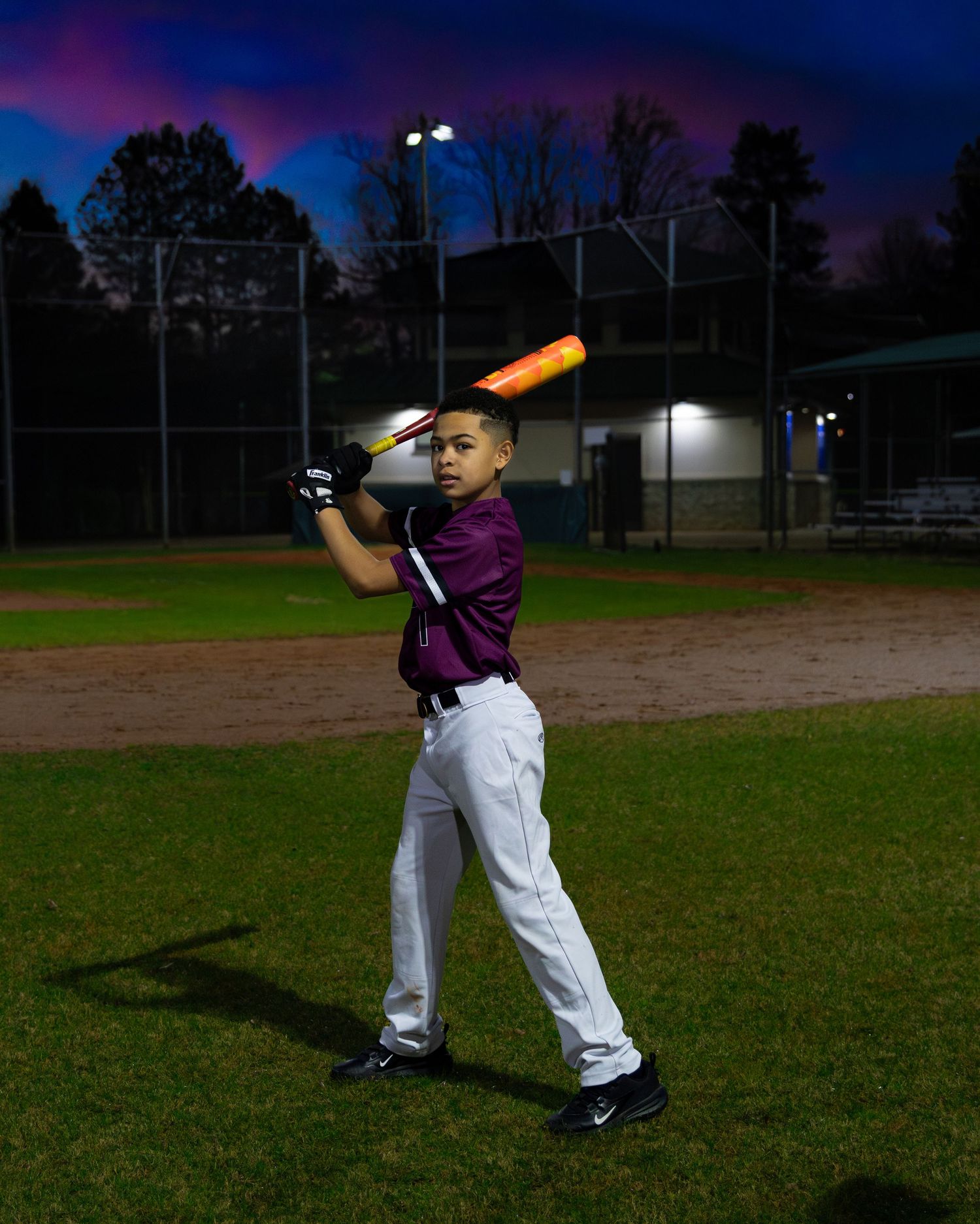 Baseball player practicing batting stance at dusk on a field with stadium lights.
