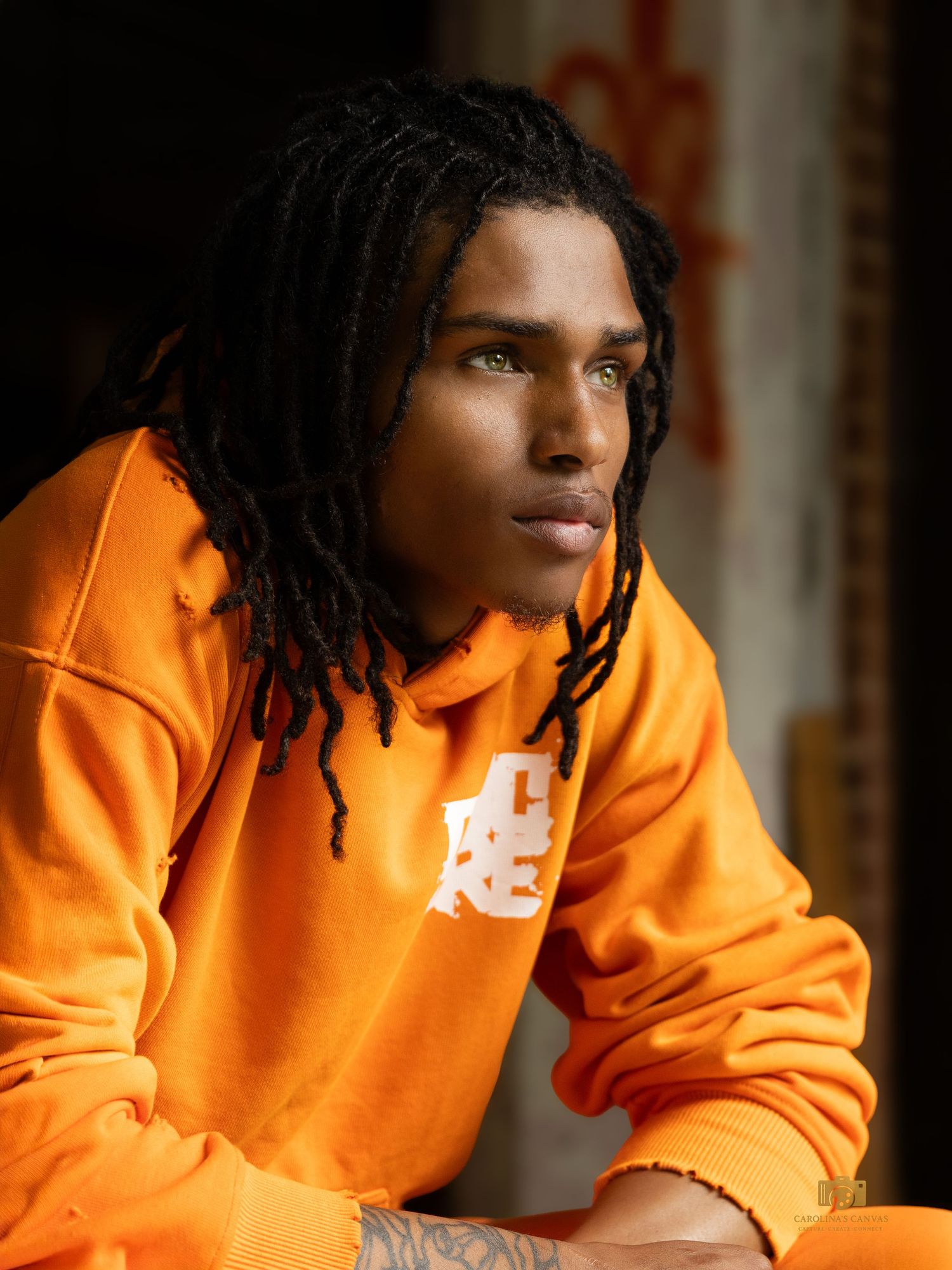 Young man with dreadlocks wearing an orange hoodie sweatshirt, gazing thoughtfully to the side in dramatic lighting.