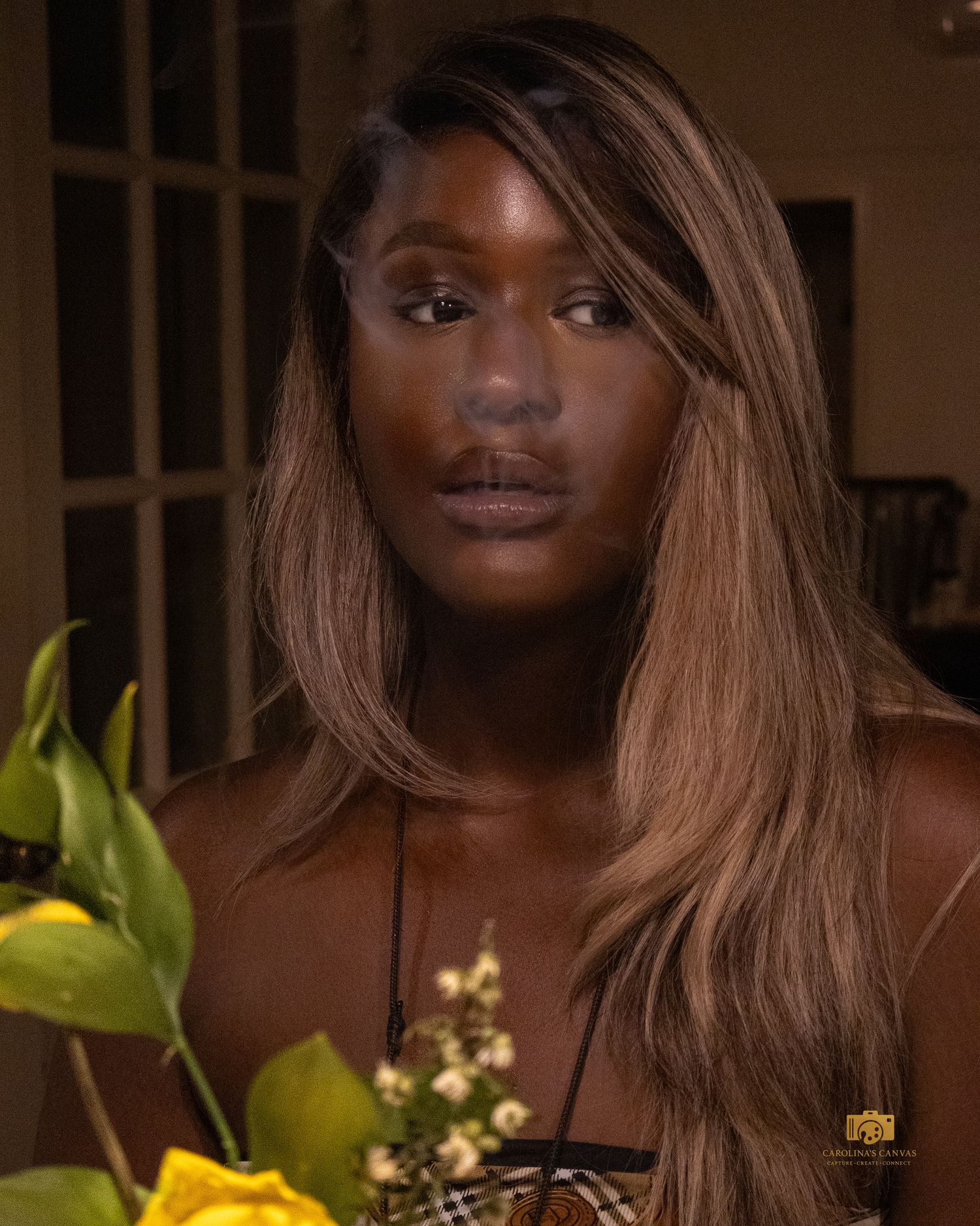 A young Black woman with long highlighted brown hair wearing a Burberry necklace, posed near yellow flowers indoors.