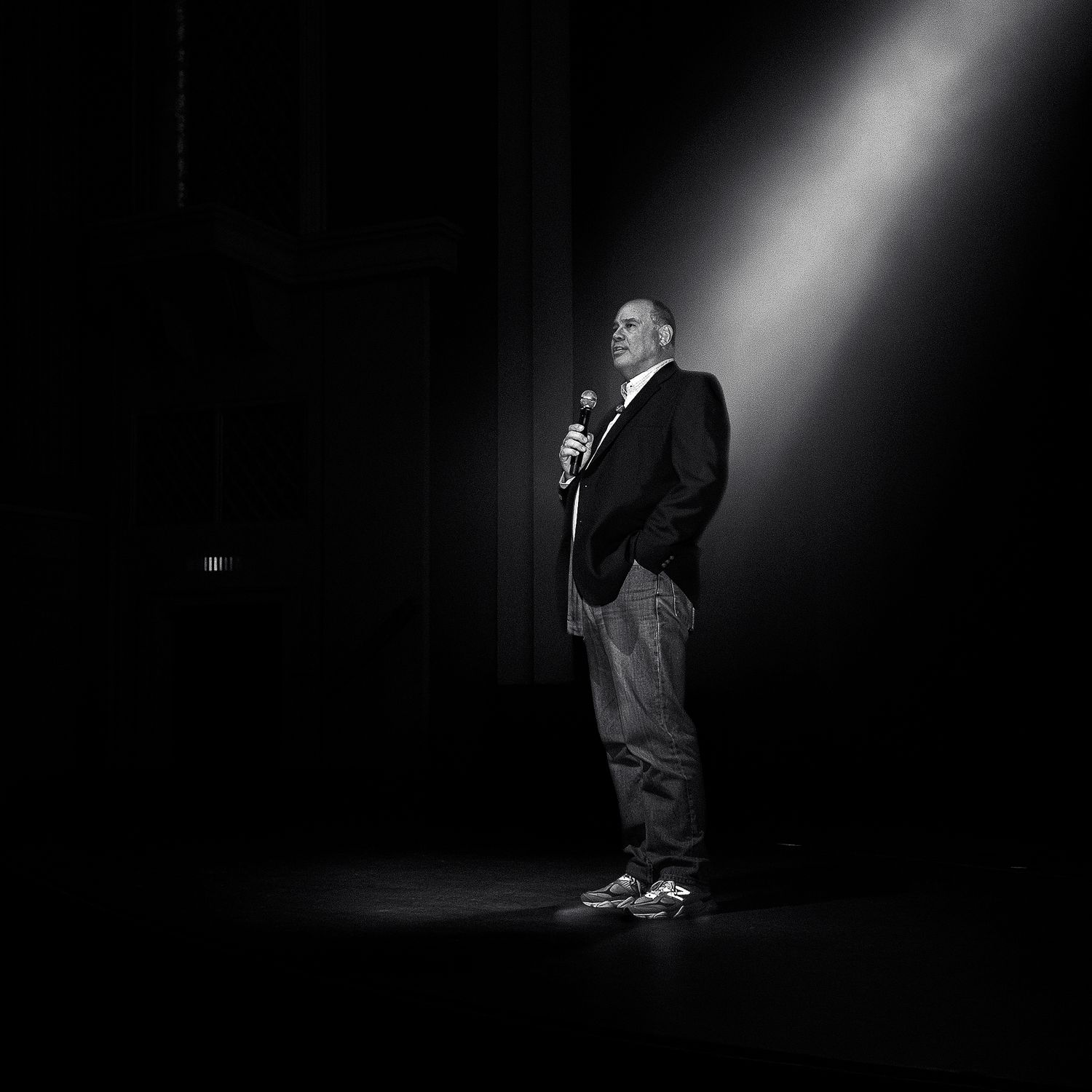 A dramatic black and white performance photo showing a person speaking on stage under a spotlight beam.