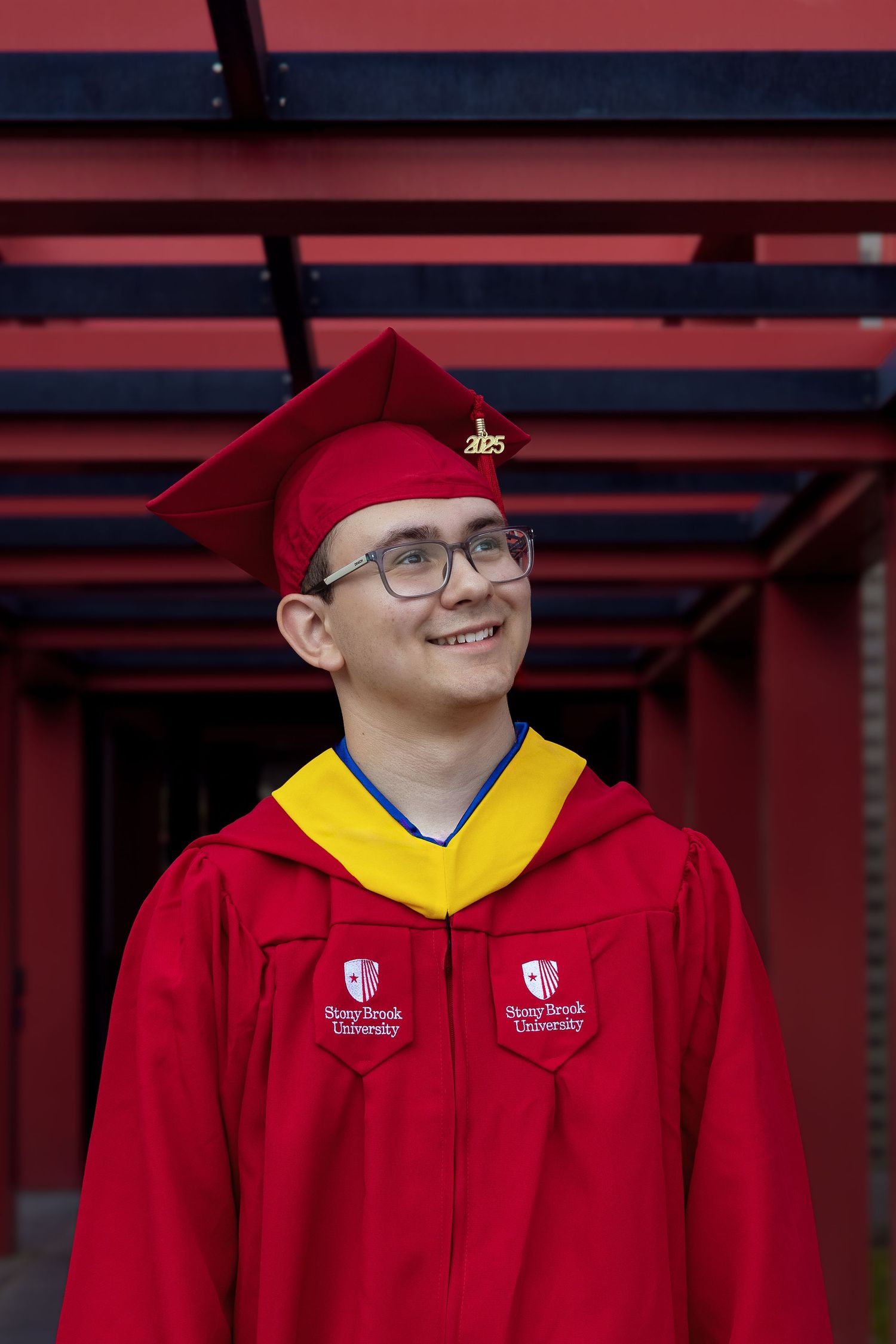 Graduate in red cap and gown with yellow hood smiles while wearing glasses under red architectural structure.