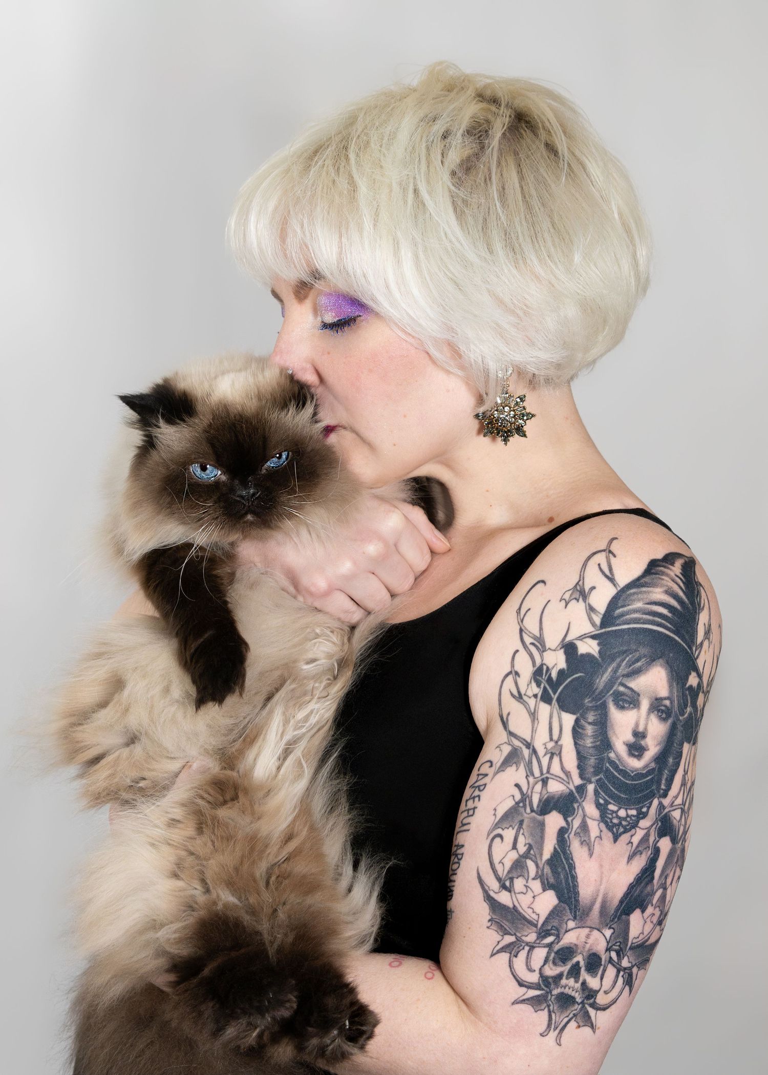 Blonde woman with purple eyeshadow and sleeve tattoo kissing a Himalayan cat against gray background.
