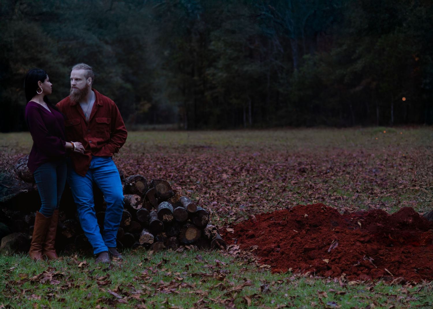 Couple standing near a woodpile in a forest clearing at dusk.