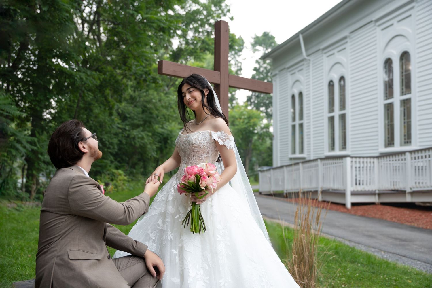 Groom kneeling before bride in white ball gown holding pink bouquet near wooden cross outside white chapel.