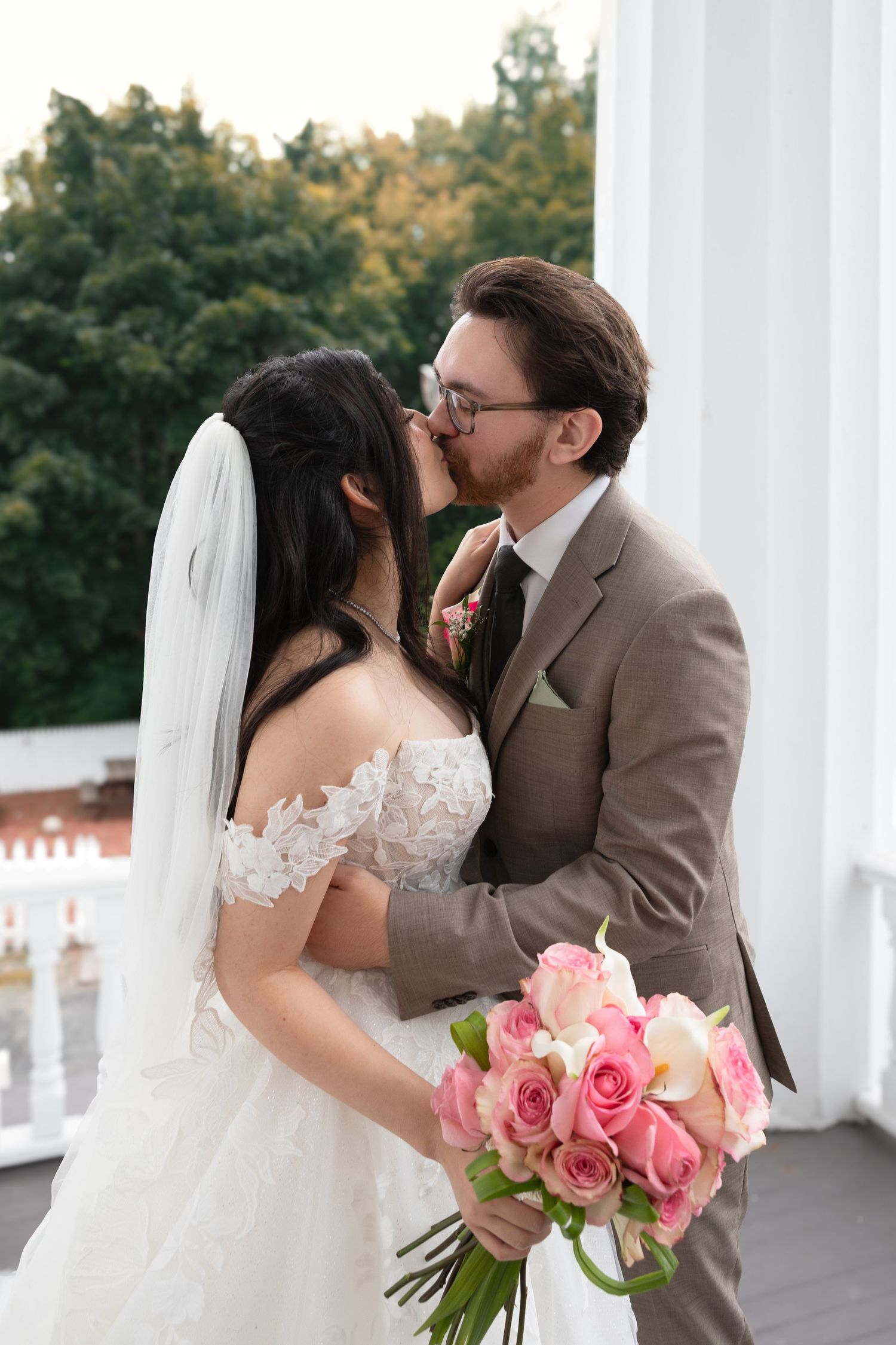 Bride and groom sharing a romantic kiss on a balcony, bride holding pink rose bouquet, wearing white lace gown and veil.