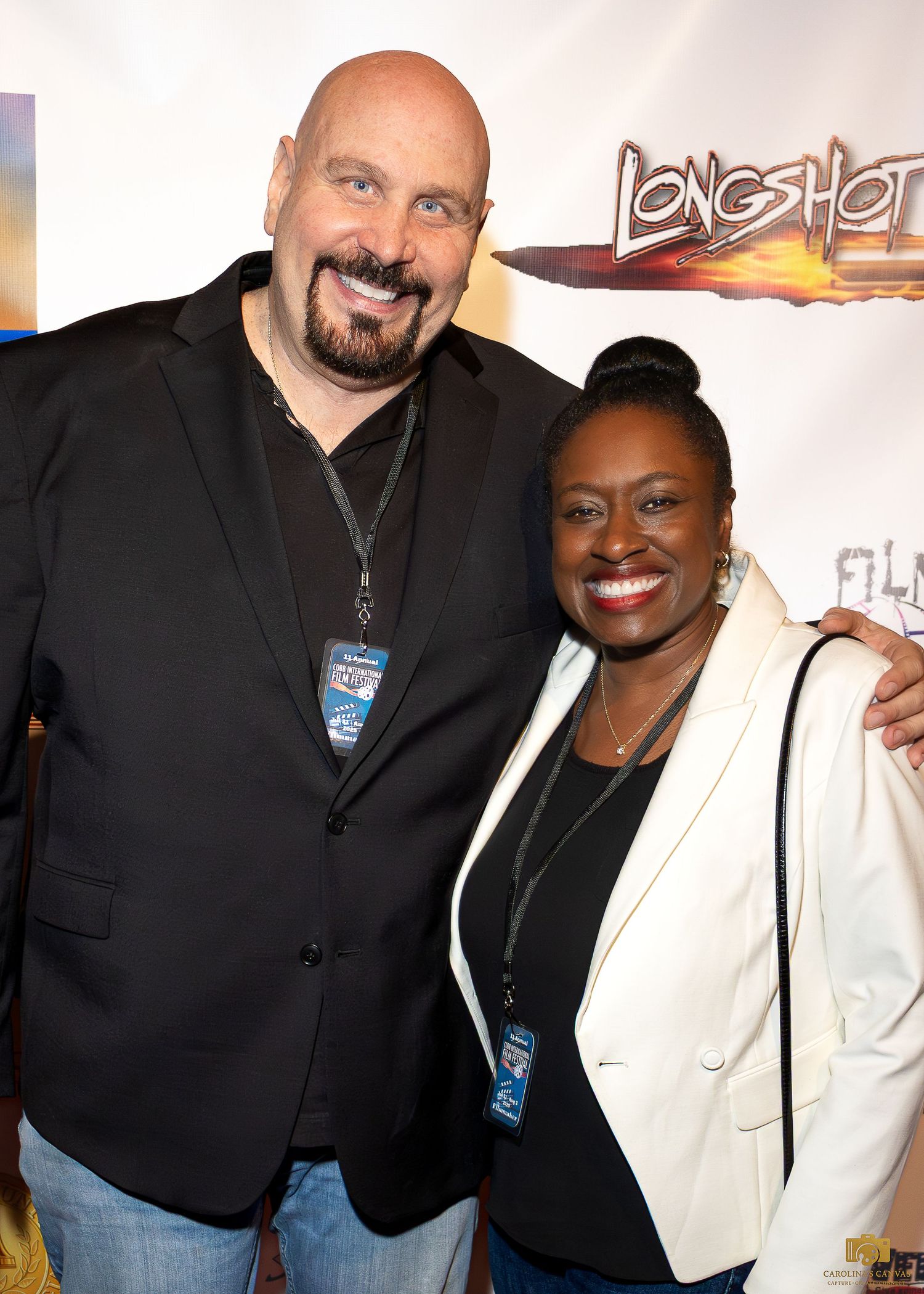 Two people wearing black and white outfits pose together at an event with a Longshot banner in the background.