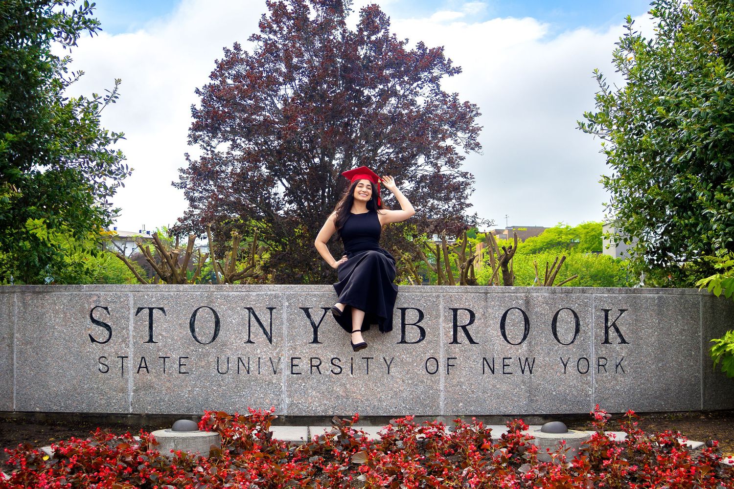 A person poses at the stone entrance sign for Stony Brook State University of New York surrounded by blooming red flowers.