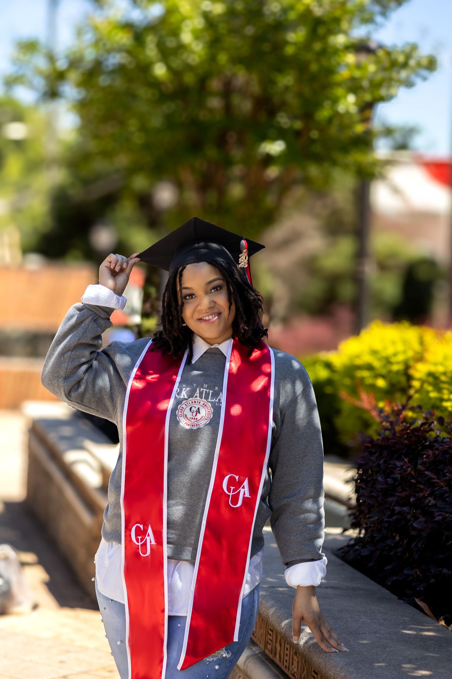 Graduation photo of student wearing a cap and gown with red stole against blurred outdoor university campus background.
