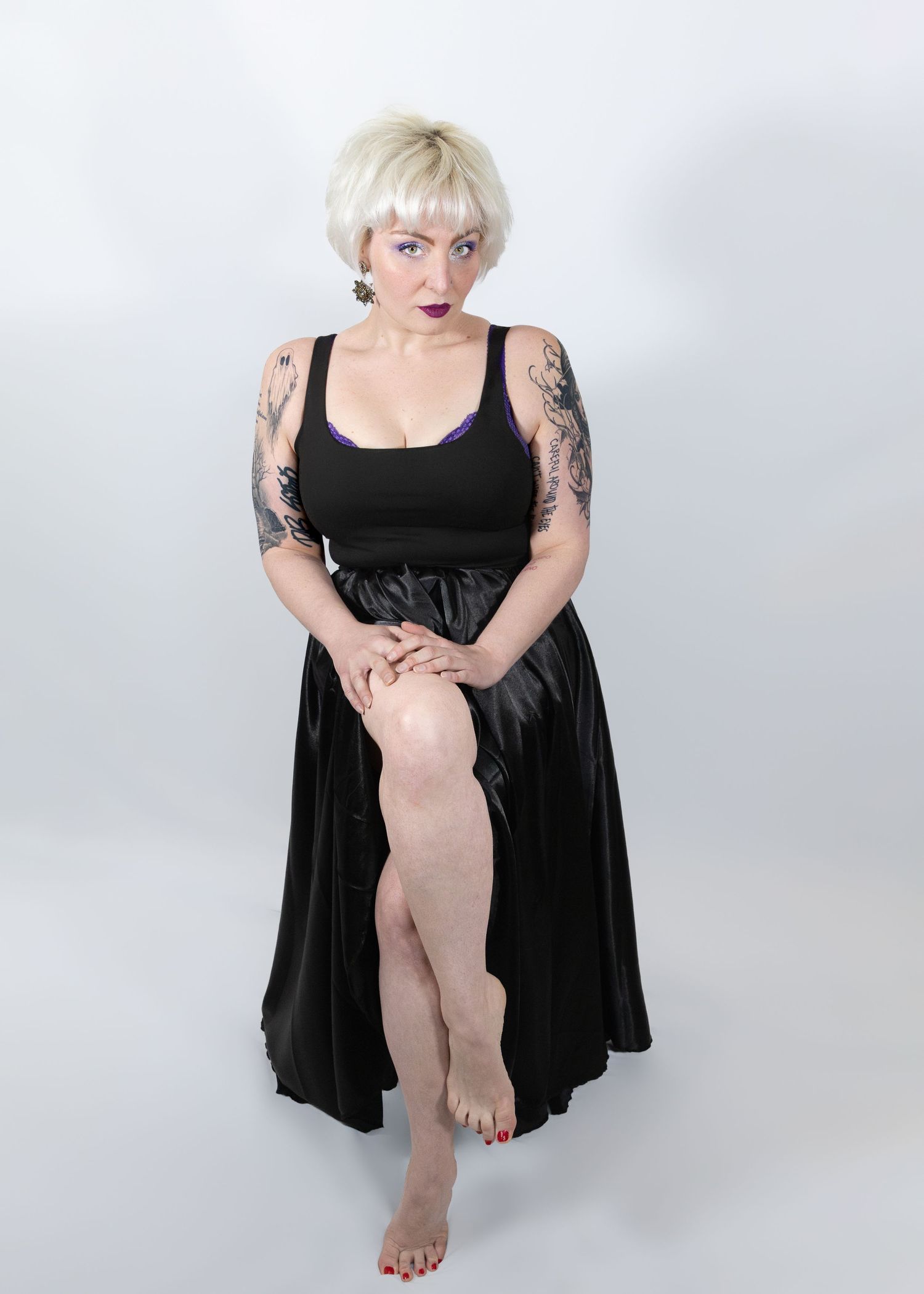 Tattooed blonde woman in black satin skirt and tank top poses barefoot against white background.