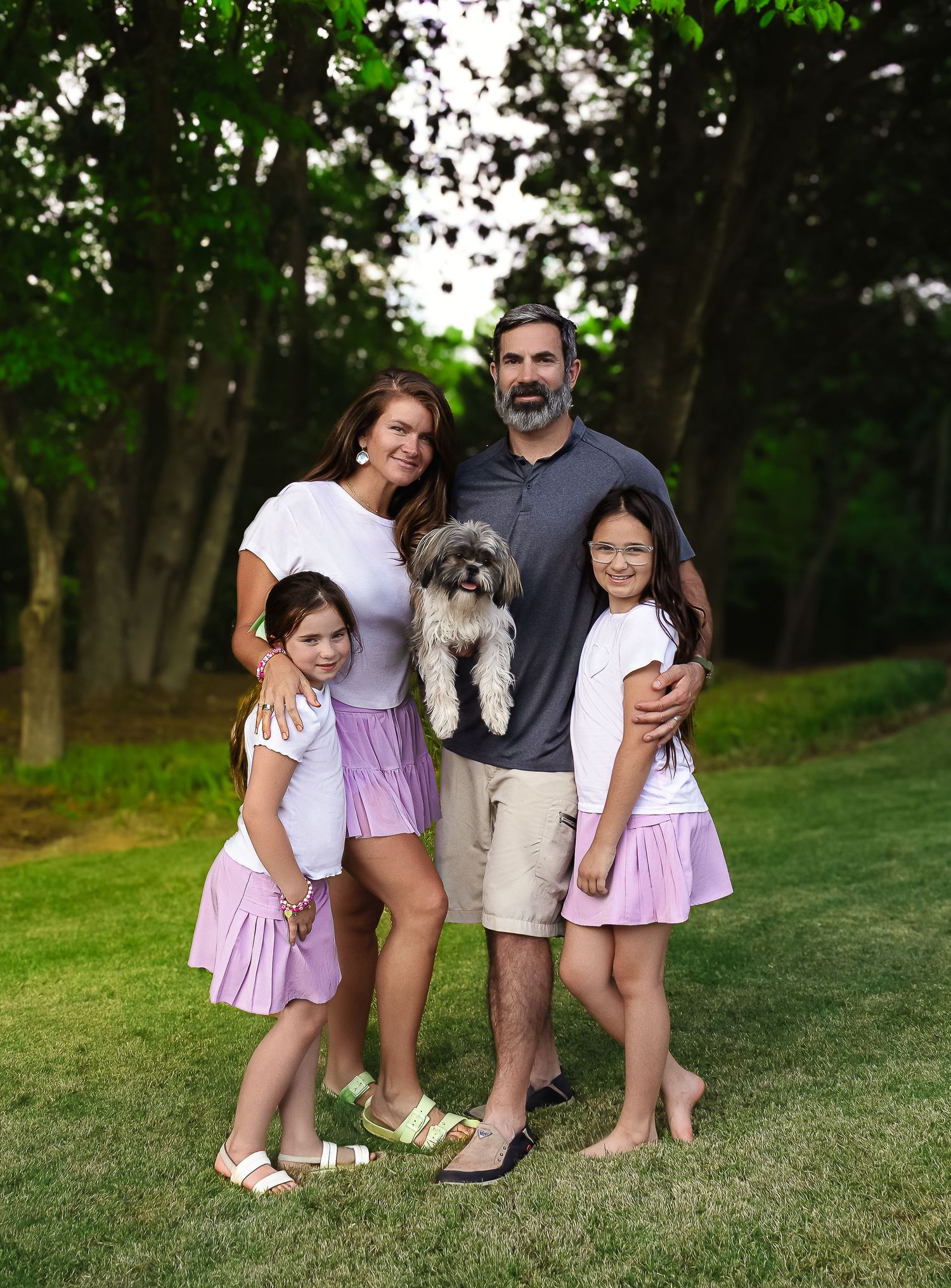 A family stands together on a grassy lawn holding their fluffy grey dog while wearing coordinated pink and white outfits.