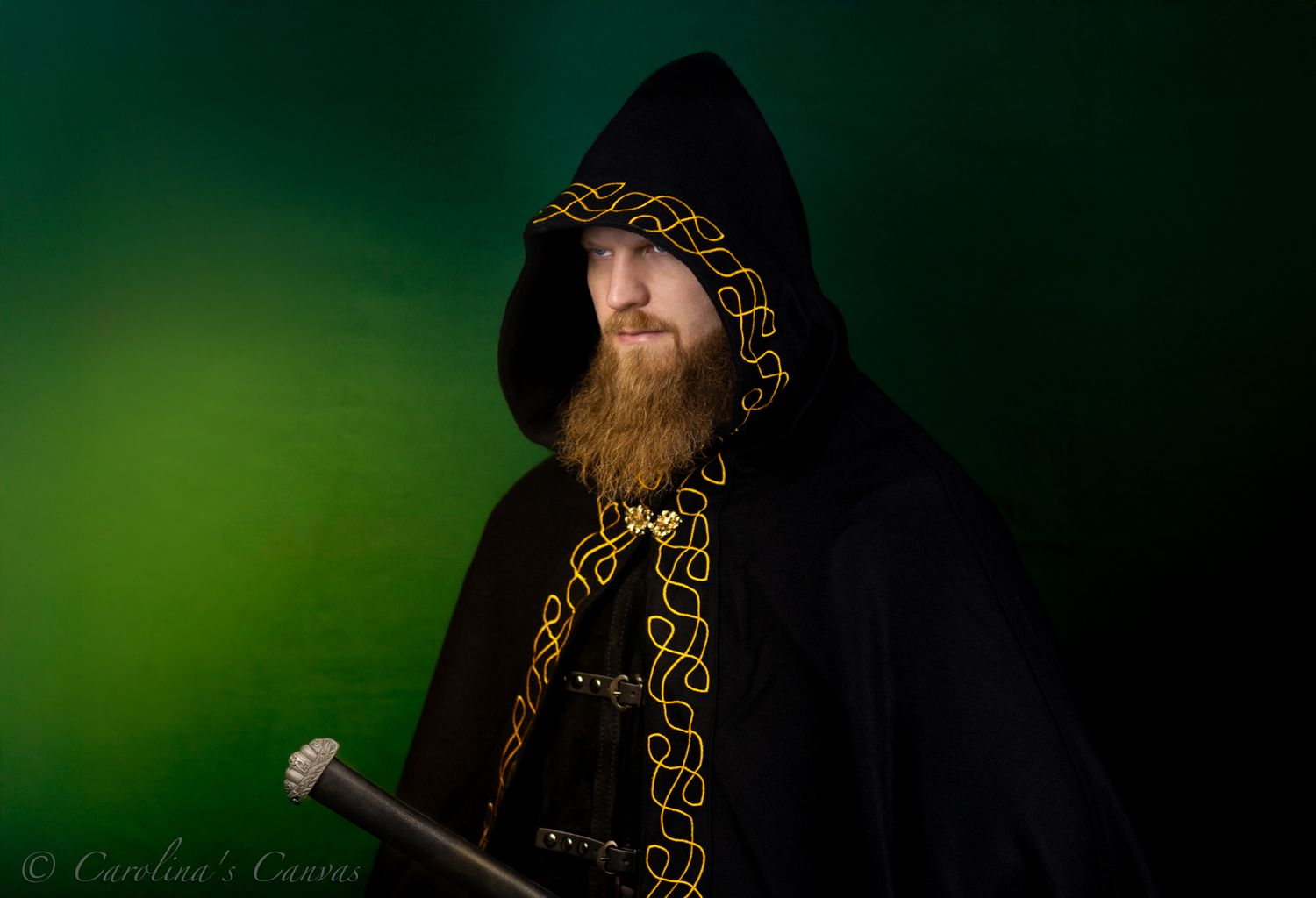 Mystical figure in black hooded cloak with golden Celtic trim stands against ethereal green background holding staff.
