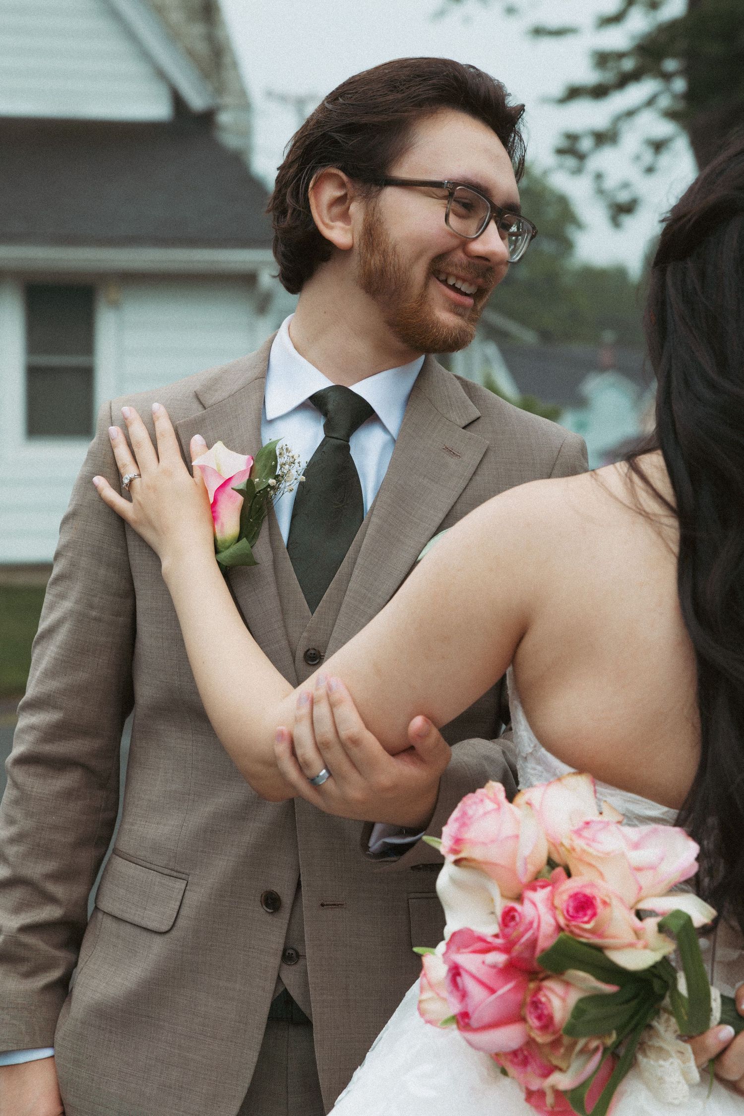 Smiling groom in tan suit with pink boutonniere embraces bride holding pink roses bouquet outdoors.