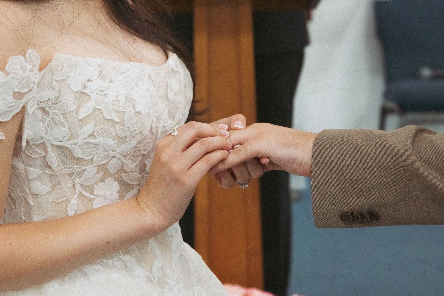 Groom placing a wedding ring on bride's finger during ceremony, bride wearing elegant lace wedding dress.