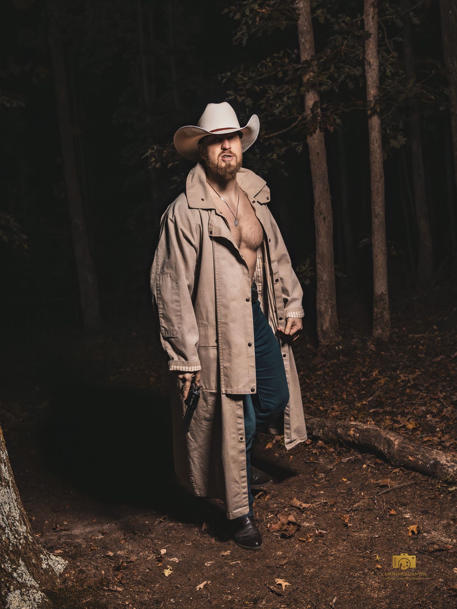 Figure in a beige duster coat and cowboy hat stands on a dark forest path during autumn.