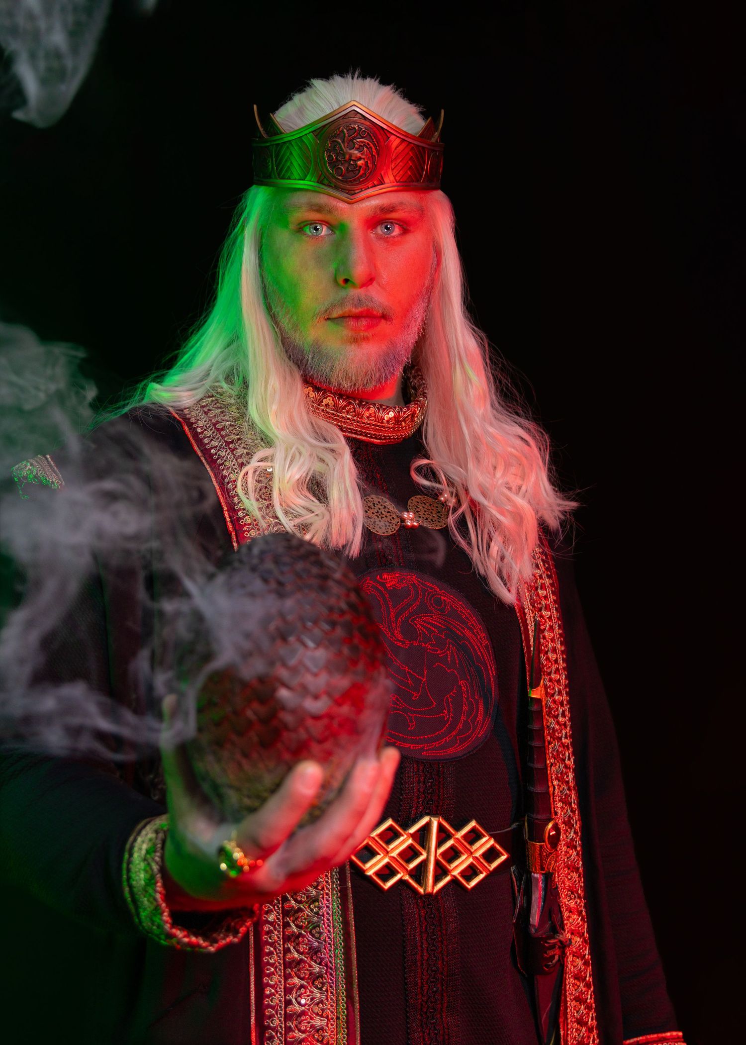 Man in medieval fantasy costume with white hair, red crown, and dark robes holds a smoking orb under green and red lights.