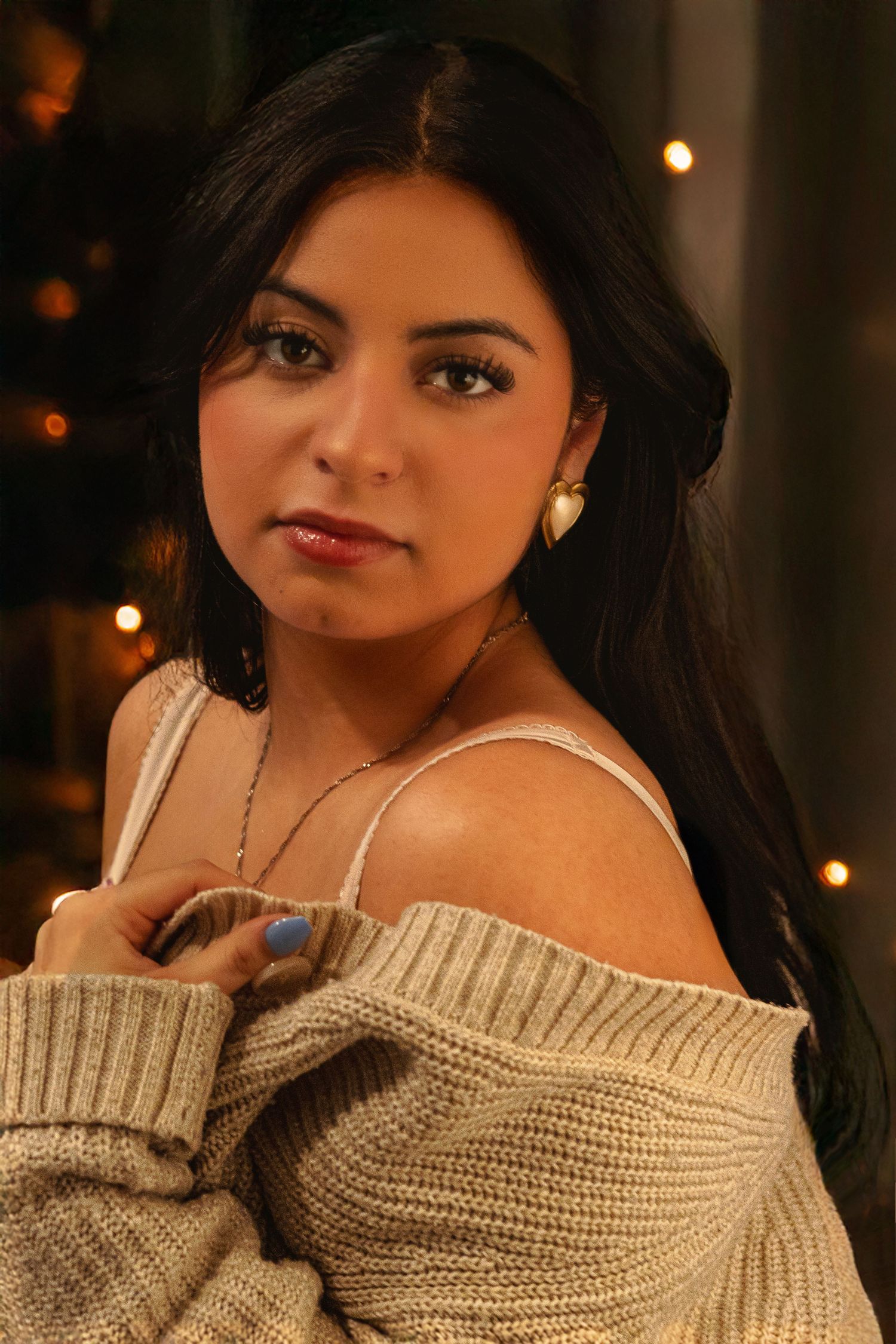 Portrait of a dark-haired woman in a beige off-shoulder sweater with moody lighting and blurred twinkle lights in background.