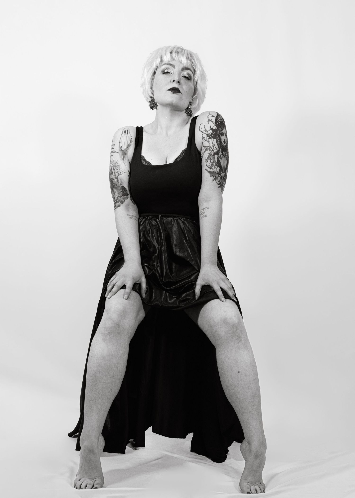 Black and white photo of tattooed woman in black dress posing confidently in studio setting.