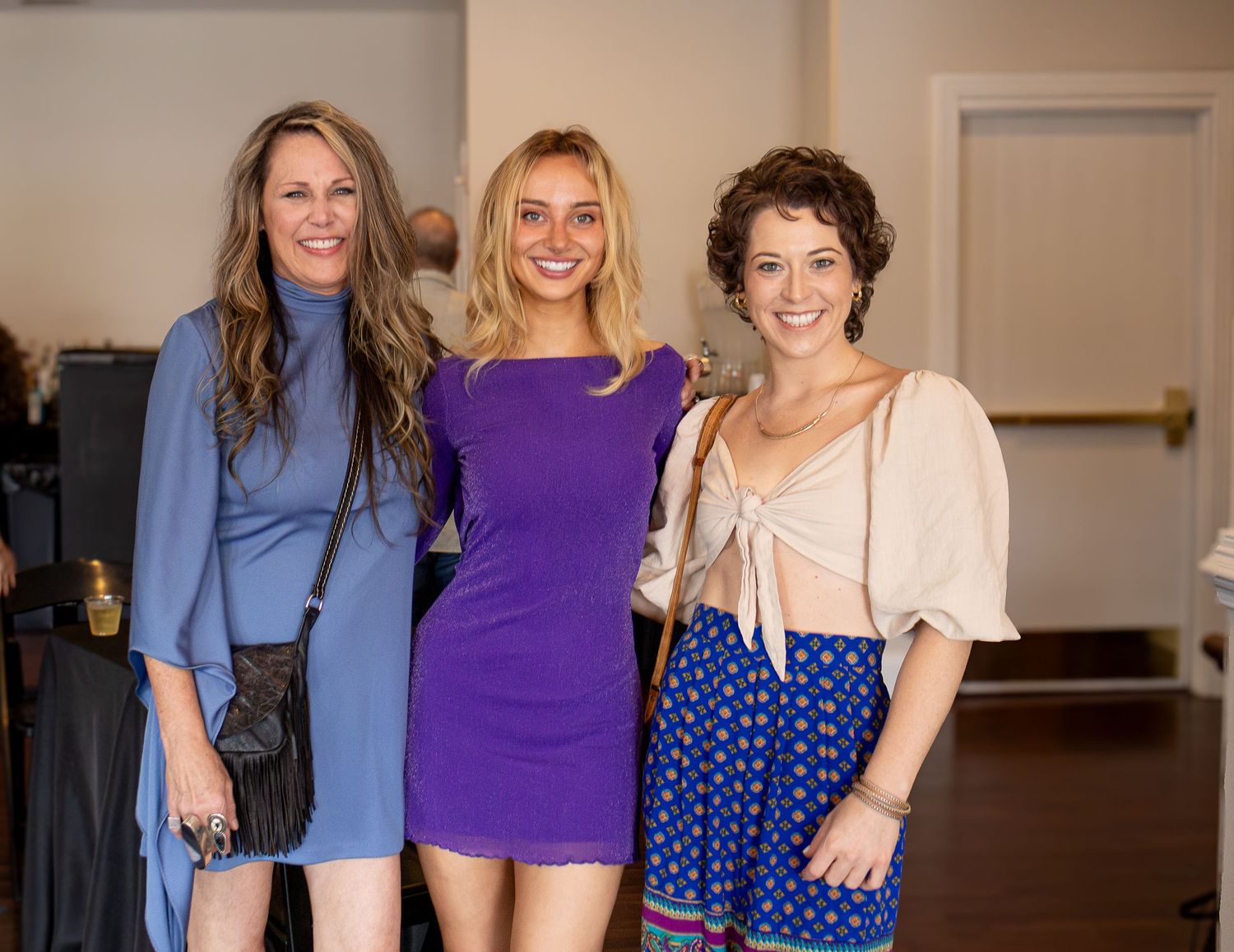 Three women pose together at an indoor event wearing a blue dress, purple dress, and polka dot dress with beige top.
