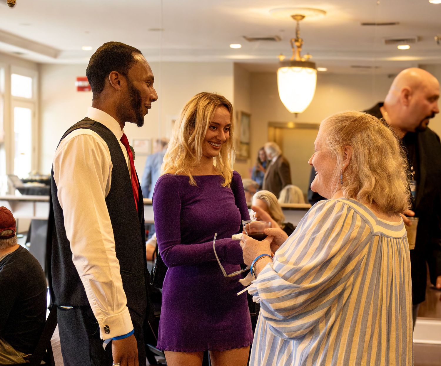 People in formal attire interact at an indoor event with elegant lighting and decor.