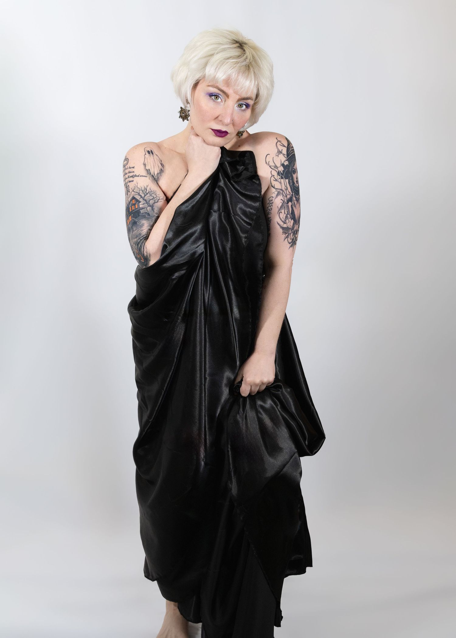 Tattooed woman with short blonde hair wearing a black satin wrap dress, posing against a white background.
