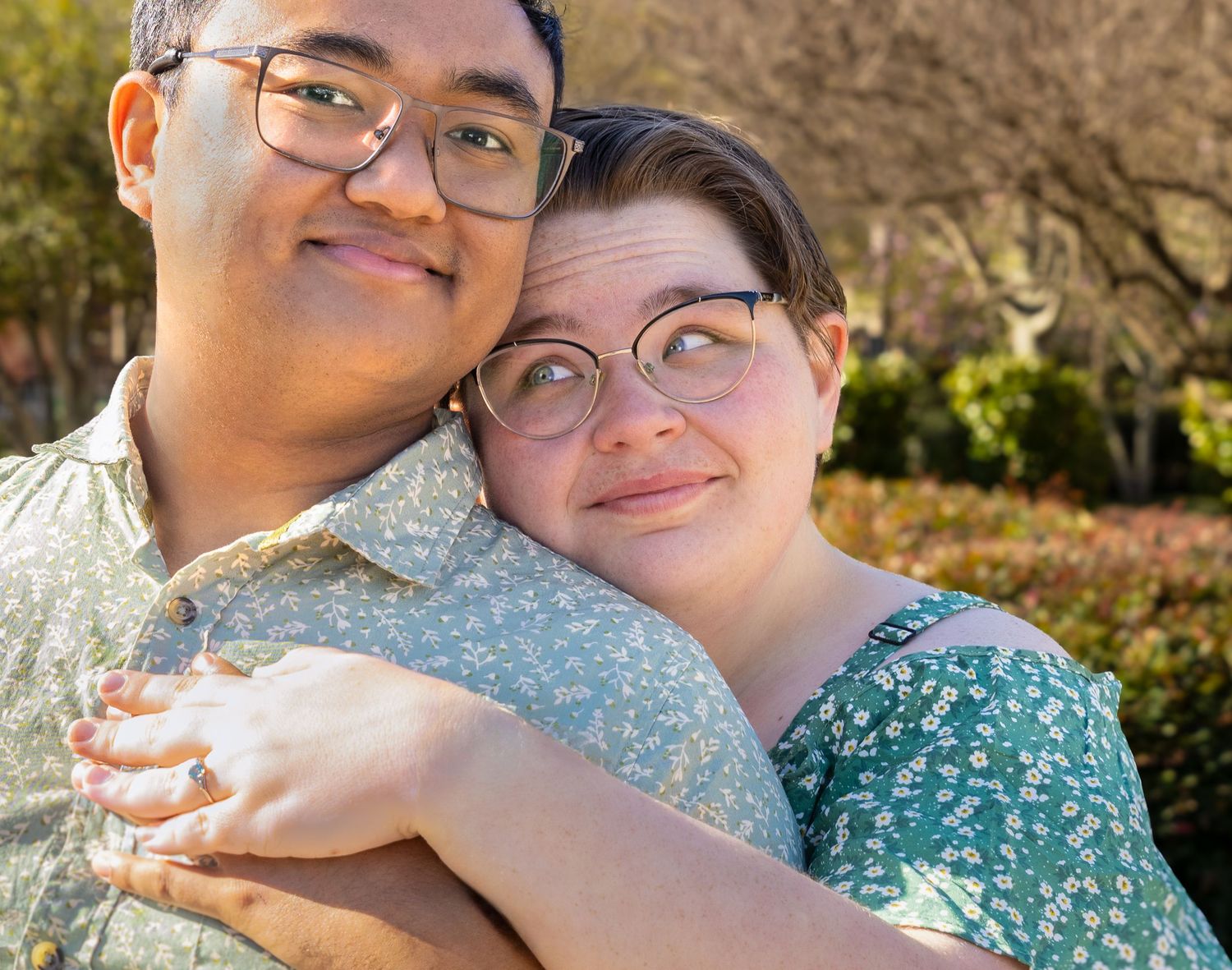 A smiling couple in glasses share a warm embrace outdoors, wearing floral shirts in a sunny garden setting.