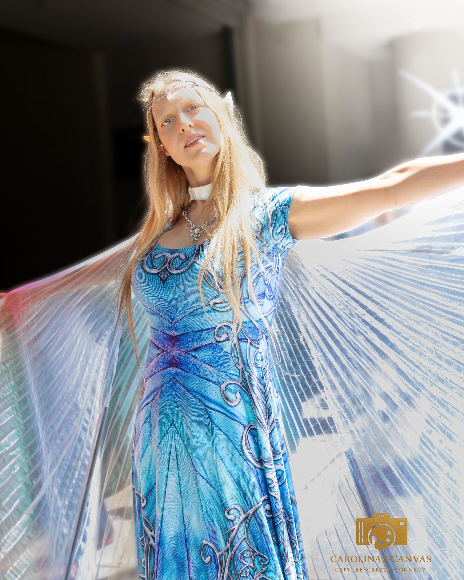 A figure in a flowing blue dress with pleated wing-like fabric panels creates an ethereal effect against a bright background.