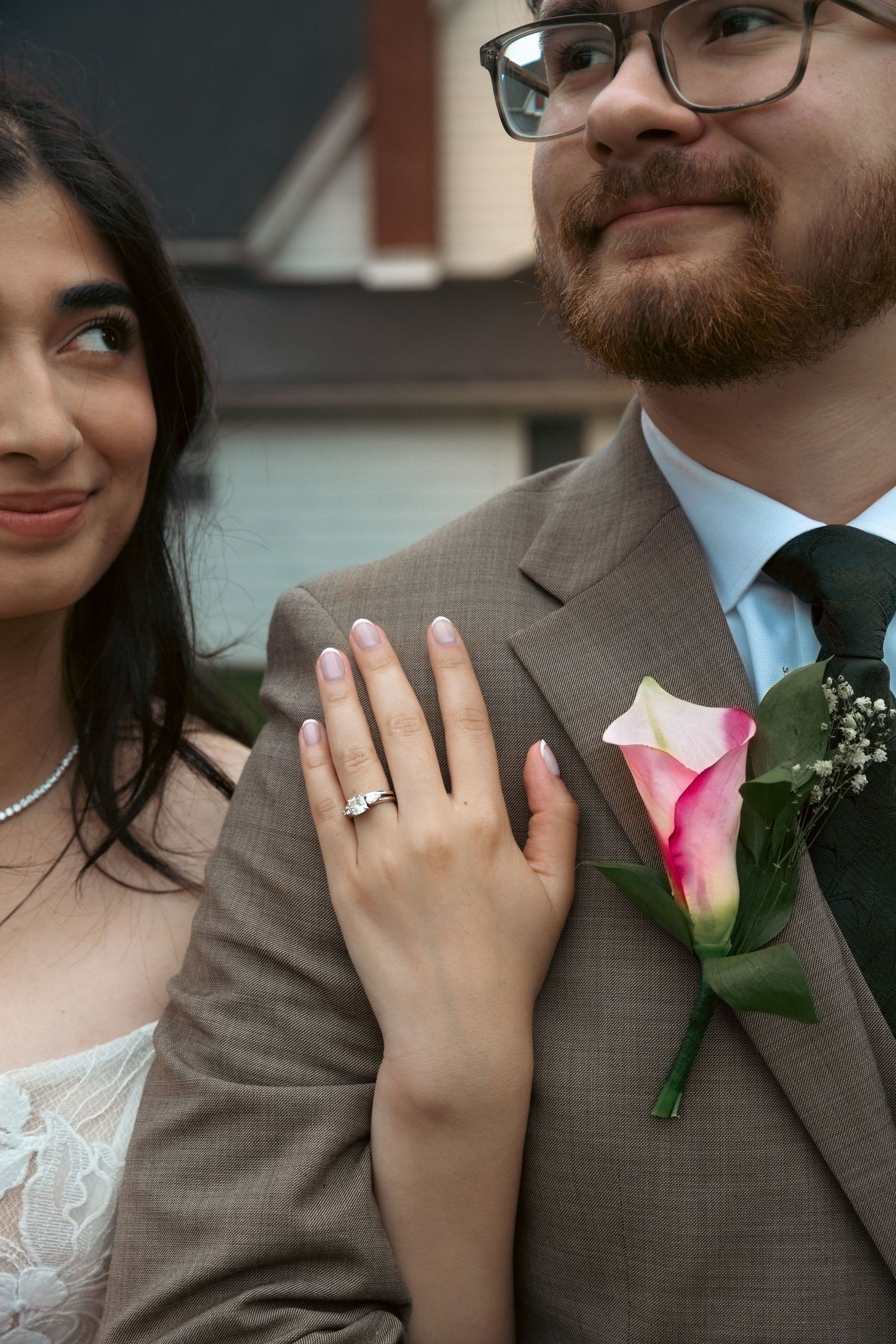 Bride rests hand on groom's brown suit lapel, showing diamond ring near pink calla lily boutonniere.