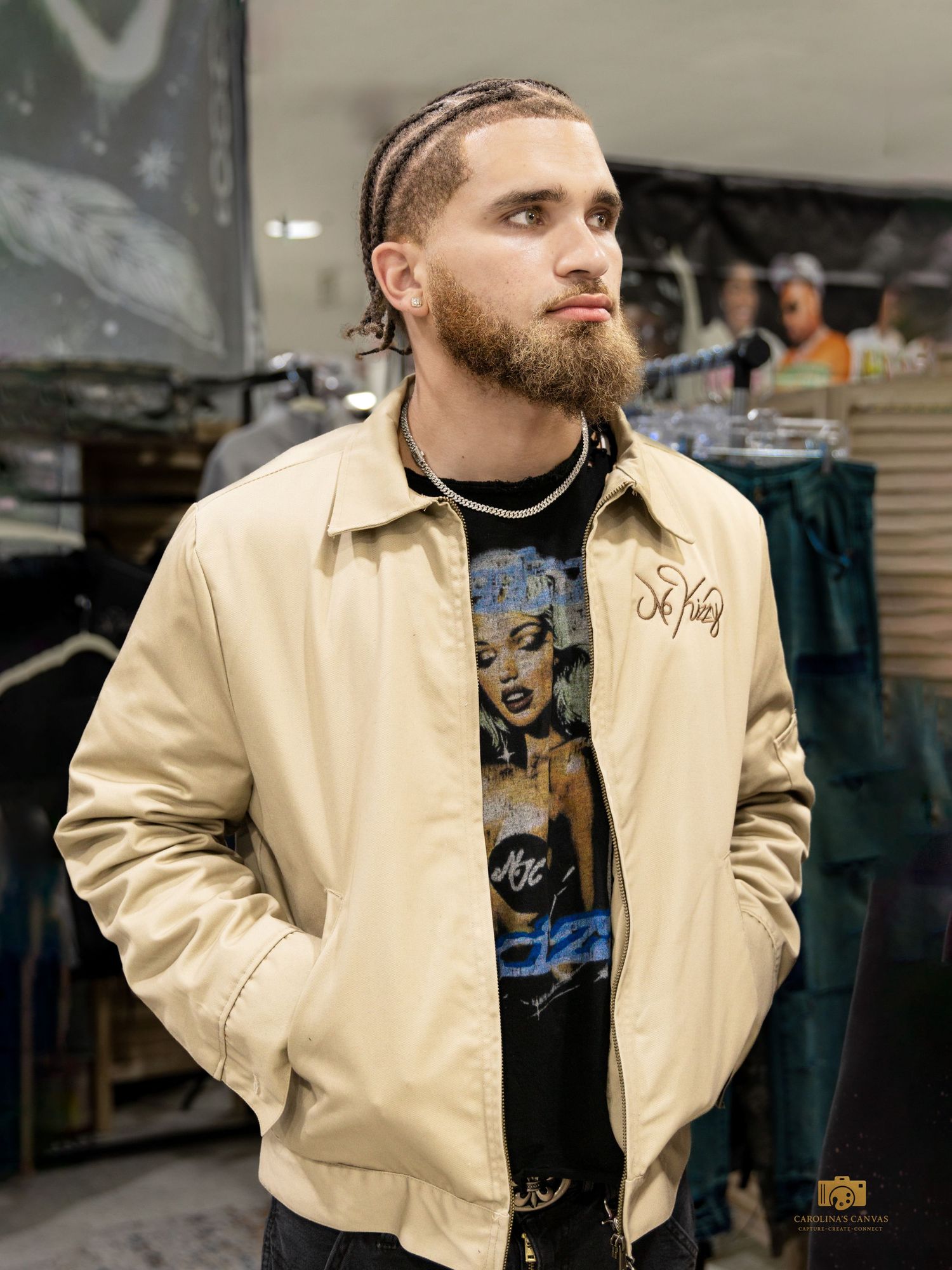 Young man with braids and beard wearing beige jacket over graphic tee in retail store.