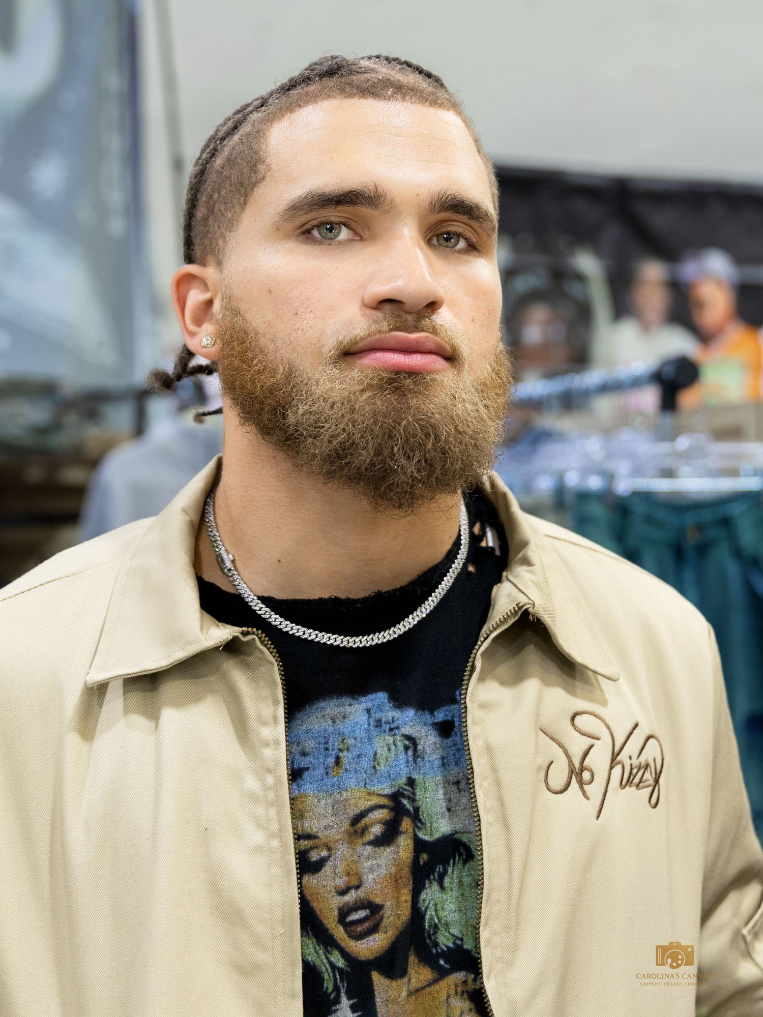 Young man with beard and chain necklace wearing beige jacket over graphic tee at retail store.