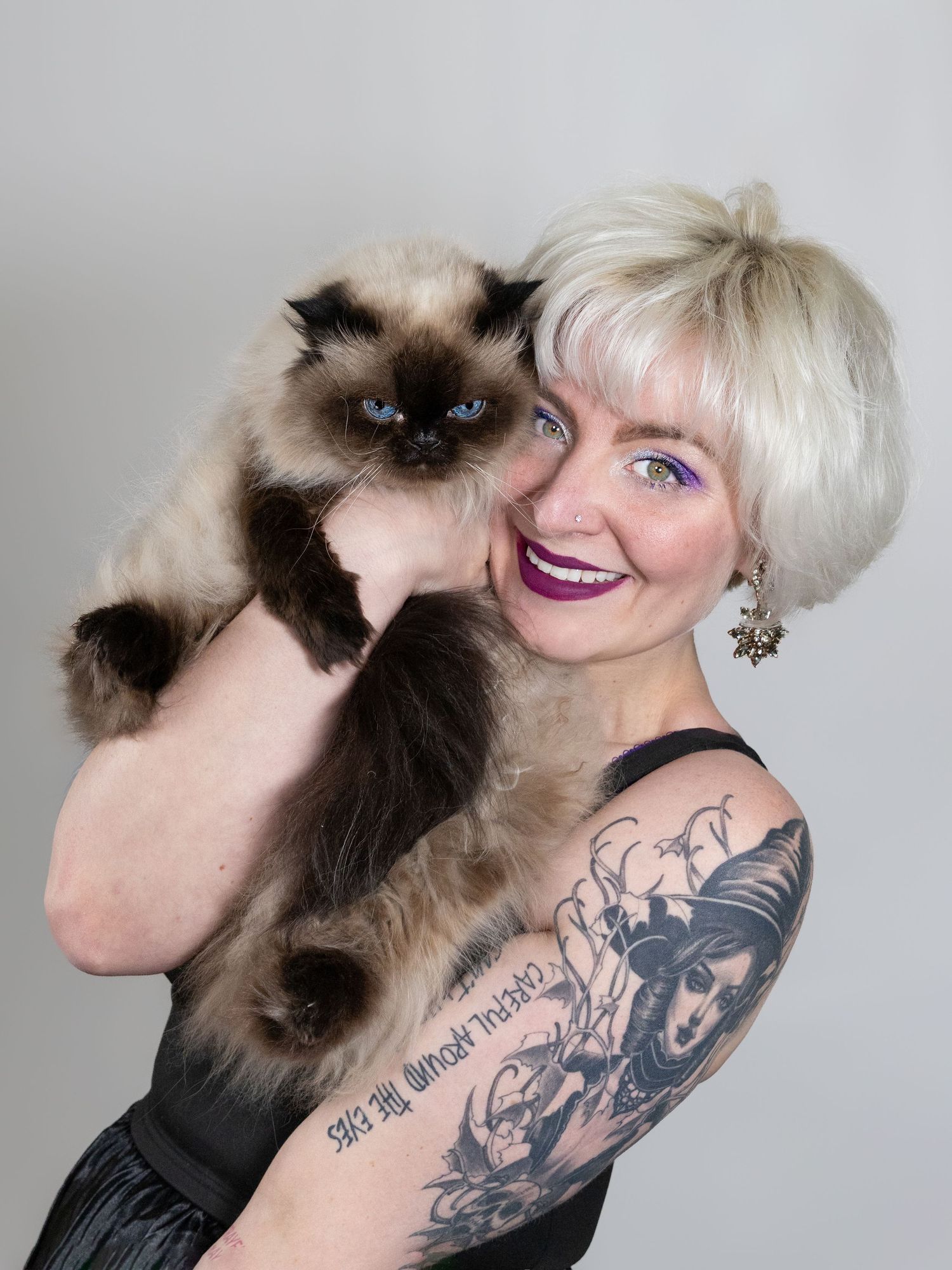 Blonde woman with tattoos and purple lipstick holds a Himalayan cat on her shoulder against a gray background.