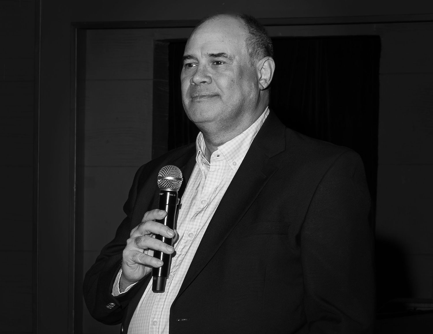 A black and white collage of four similar images showing someone speaking into a microphone at a formal event.
