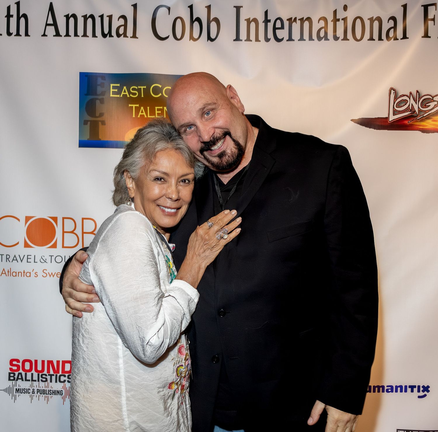 A pair poses warmly together at the Cobb International Film Festival with event logos behind them.
