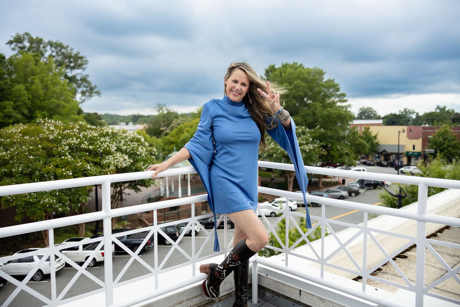 A person in a blue sweater dress and boots poses on a white deck railing on a cloudy day.