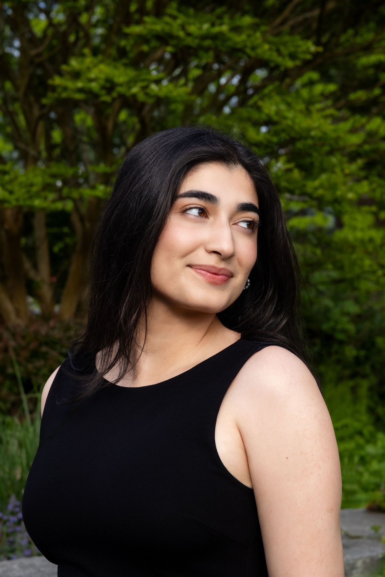 A woman in a black tank top smiles while standing in front of lush green foliage on a sunny day.