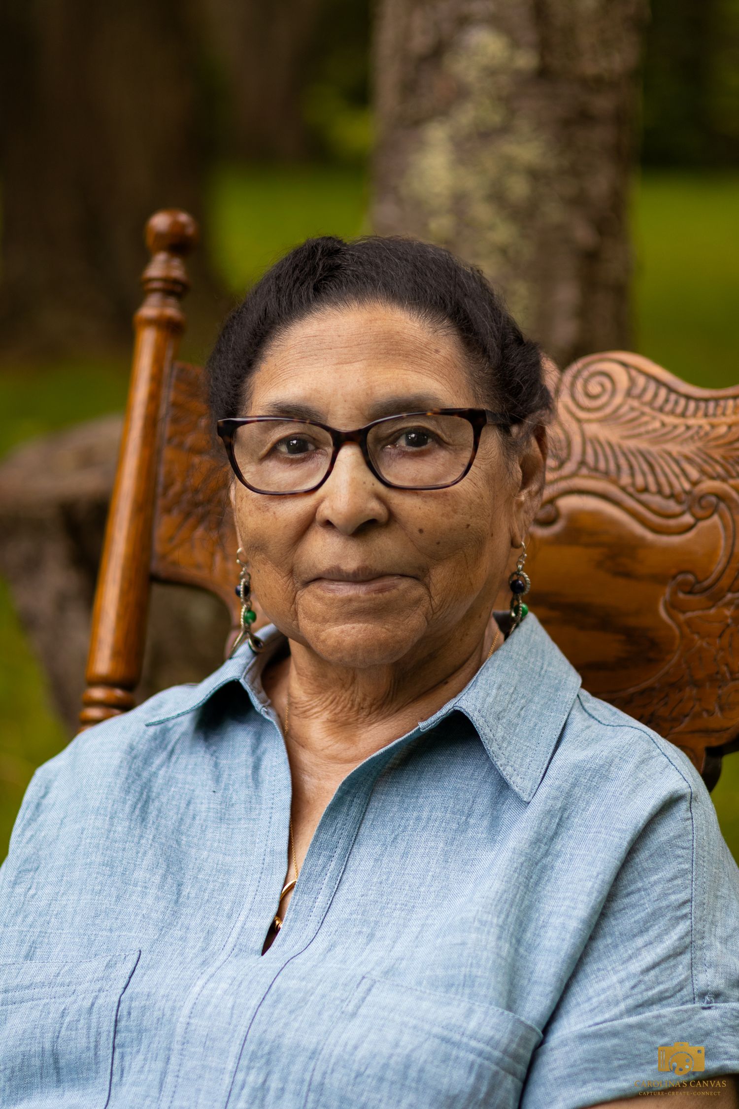 An elder wearing glasses and a light blue linen shirt sits in an ornate wooden chair outdoors.