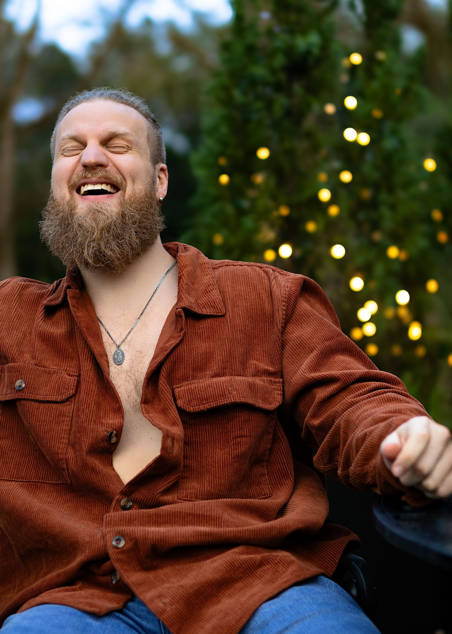 Person with beard laughs while wearing brown button-up shirt against holiday lights.