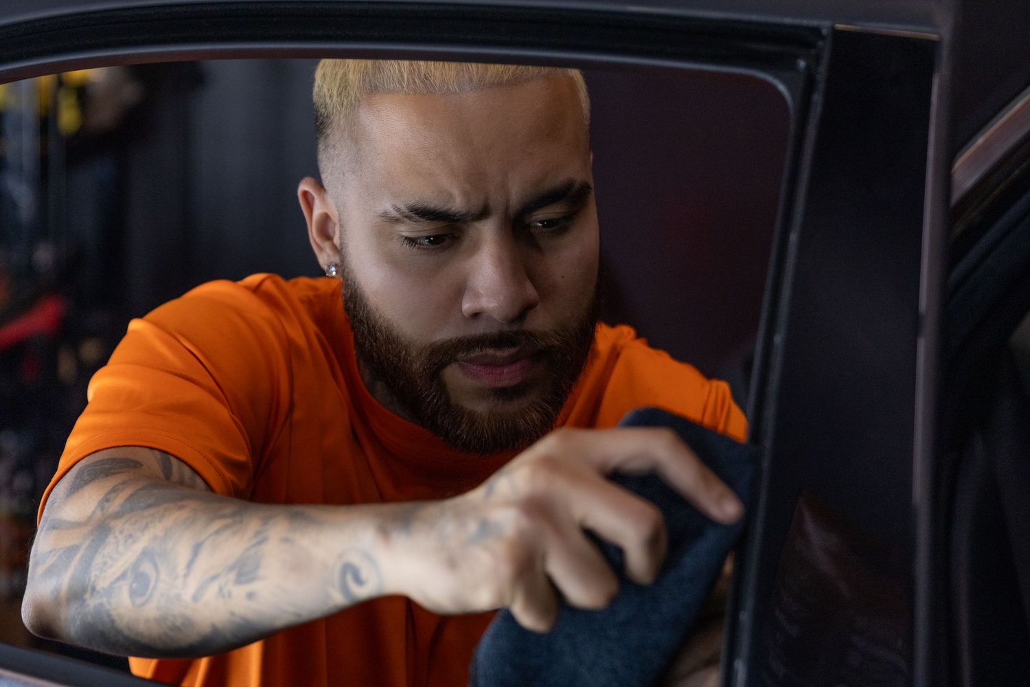 Tattooed man in orange shirt carefully cleaning car window with microfiber cloth in auto detailing shop.
