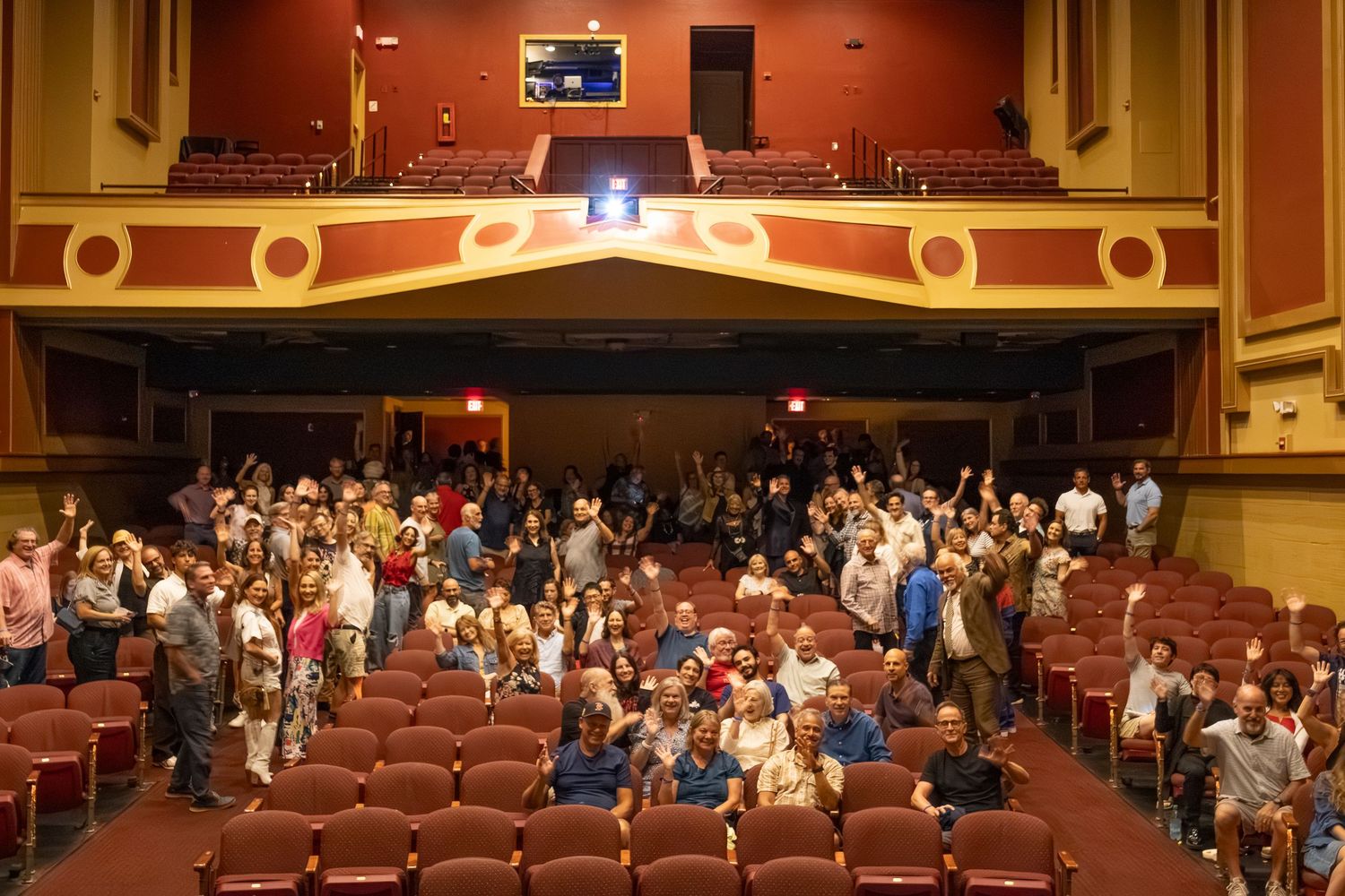 Large theater auditorium with rows of red seats and audience members scattered throughout.