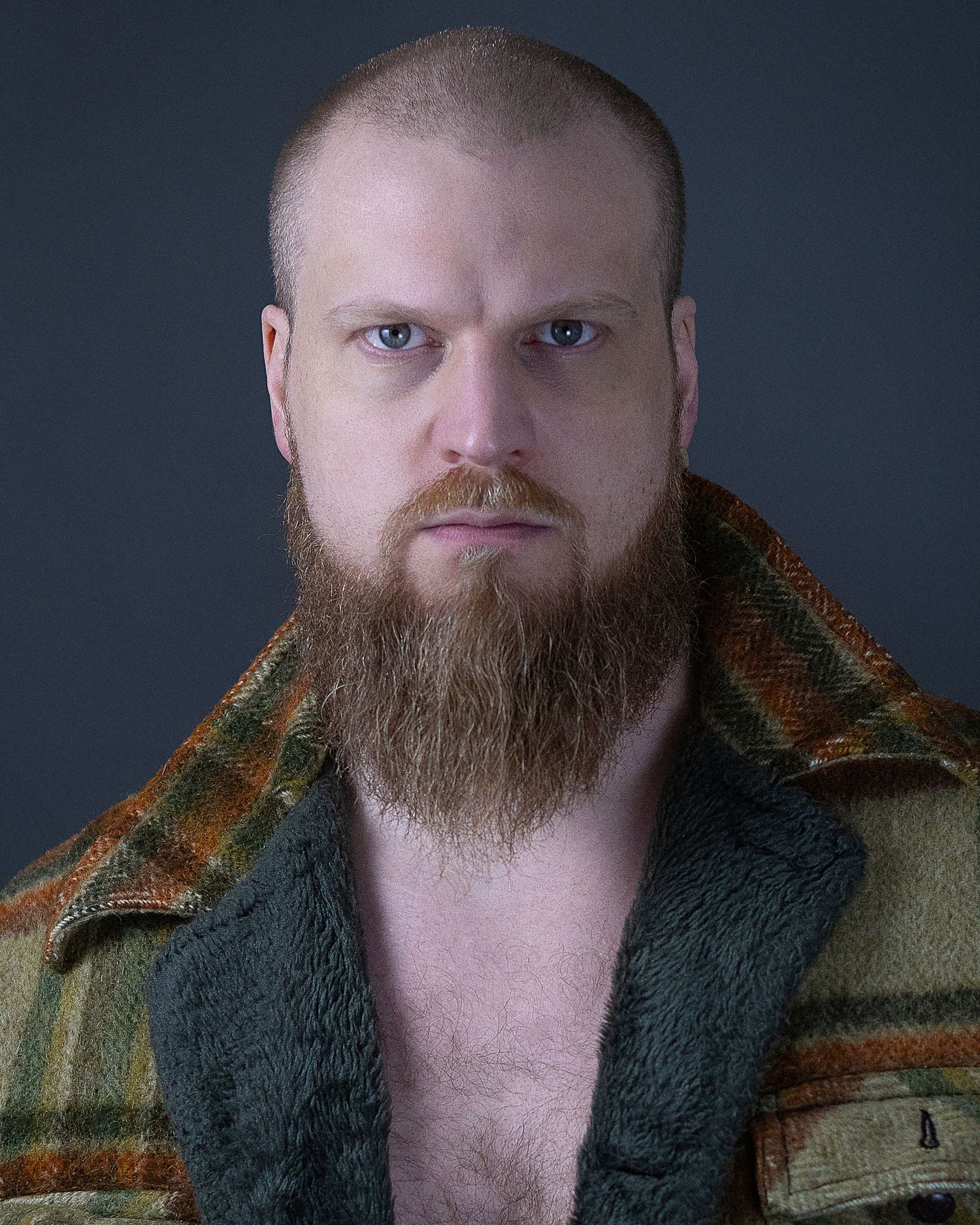Portrait of someone wearing a plaid flannel coat with a full brown beard against dark background.