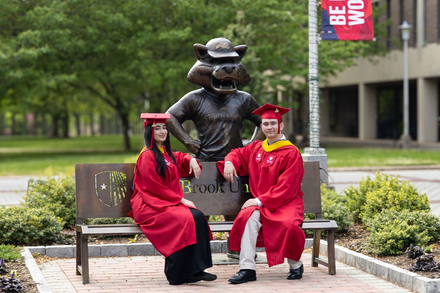 Two graduates in red caps and gowns pose beside a university mascot statue on a brick path on campus.