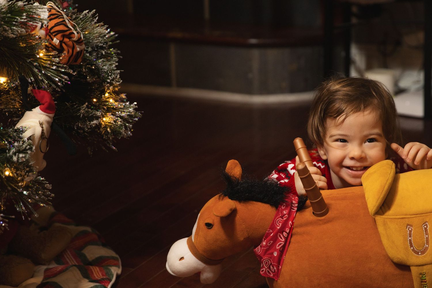 A child sits near a Christmas tree with a large plush reindeer toy wearing a red bow.