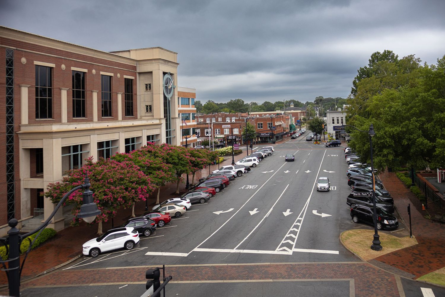 Panoramic view of a downtown street with brick buildings, parked cars and trees under cloudy skies.