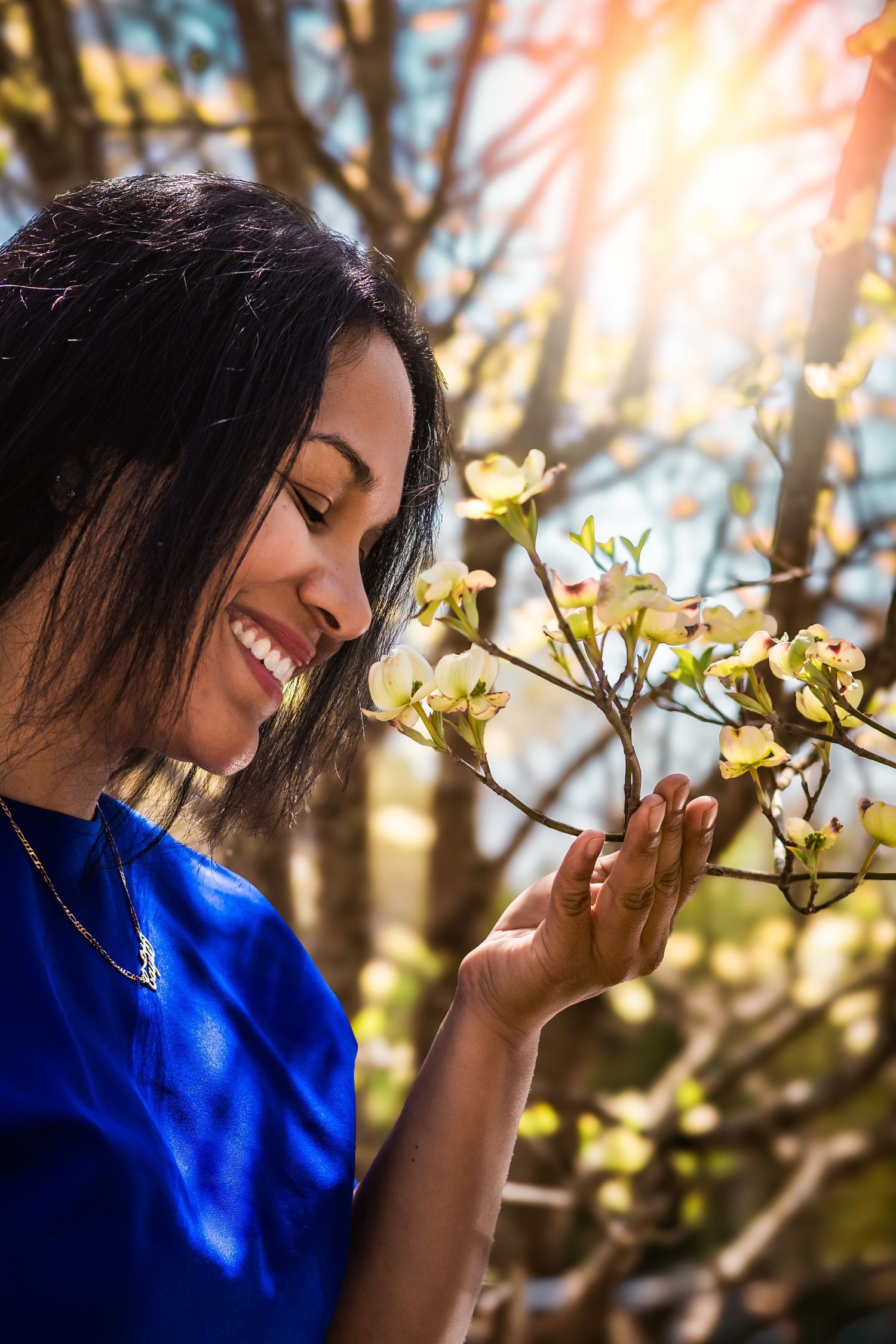Person in bright blue attire gently touches blooming flowers while smiling in warm sunlight.