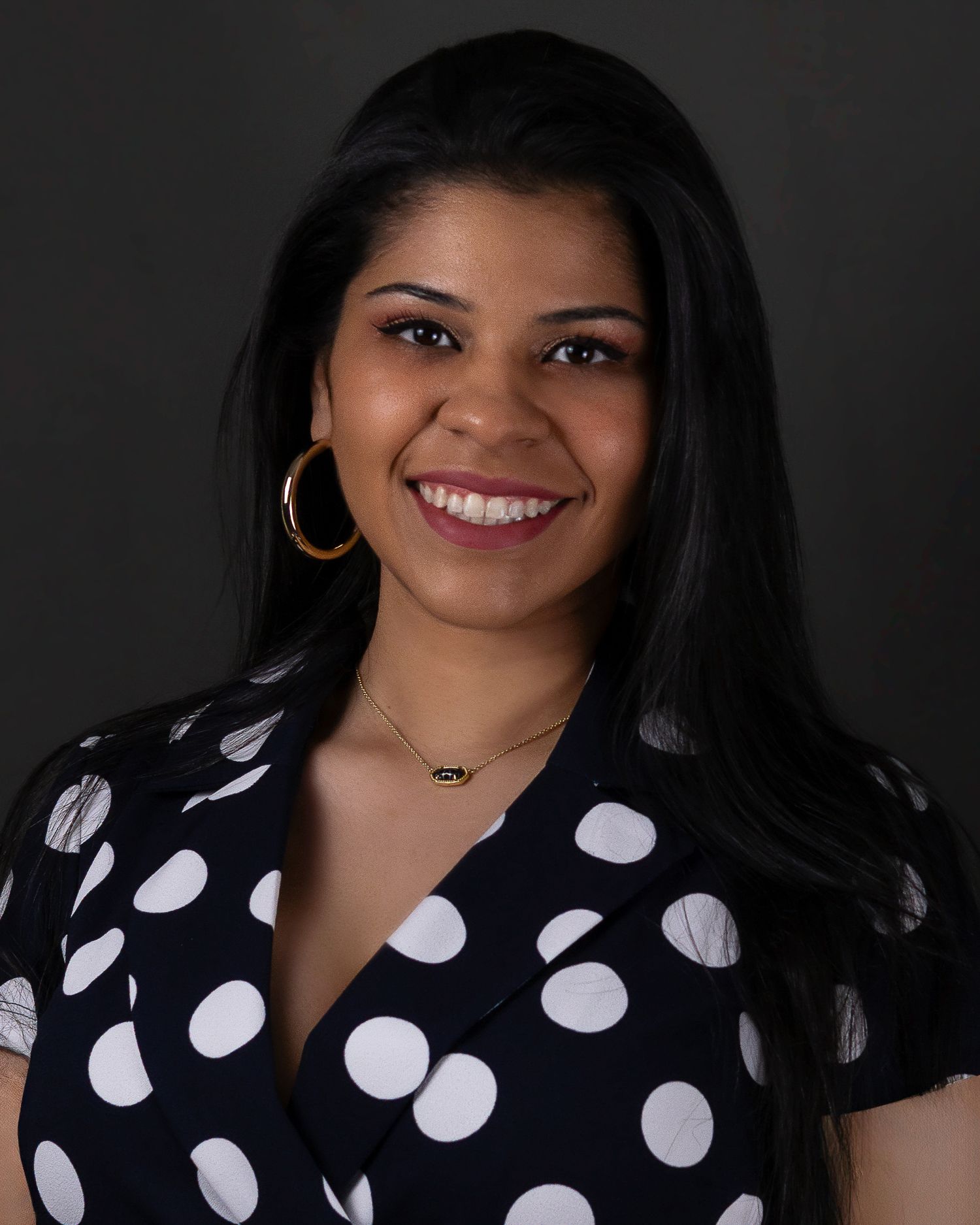 Professional headshot of a person wearing a polka dot dress and gold jewelry against a dark background, smiling brightly.