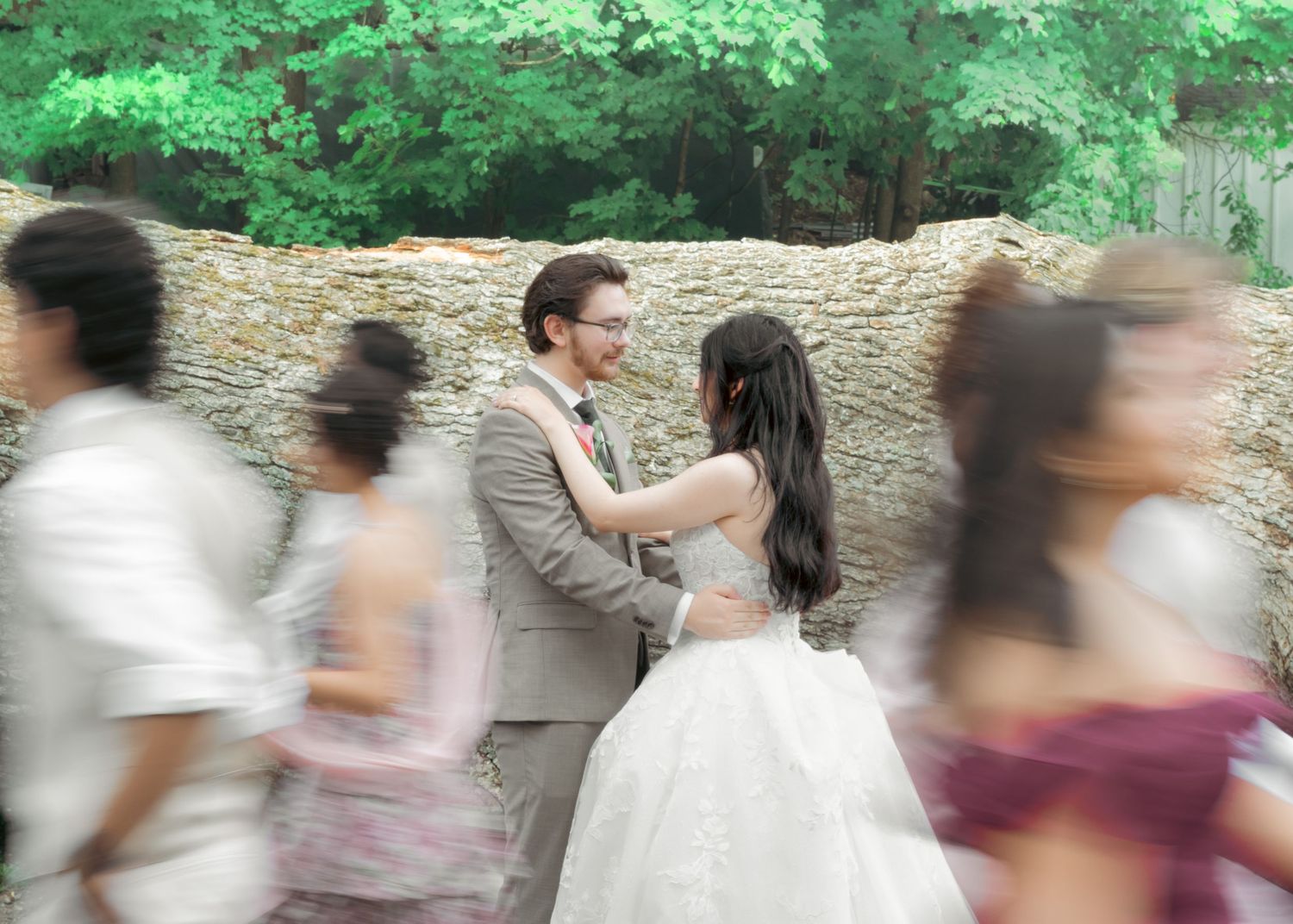 A bride and groom share a tender moment outdoors, surrounded by blurred wedding guests and lush green trees.